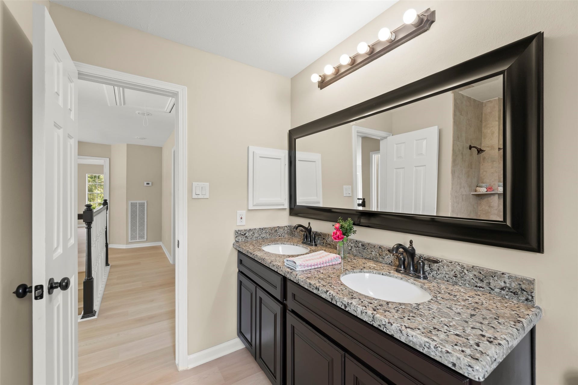 Stylish granite countertop and elegant dark wood cabinets. It includes a large mirror with ample lighting above. The neutral walls and light wood flooring create a bright and welcoming atmosphere, with a hallway visible through the open door.