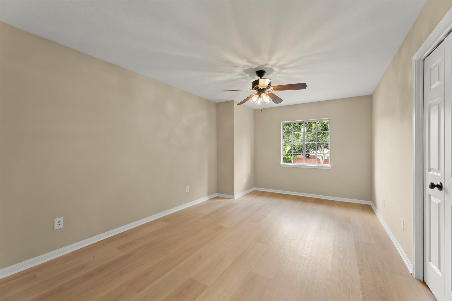 Bright, spacious room with light wood flooring, neutral walls, a ceiling fan, and a large window offering natural light. Ideal for a bedroom or office space.