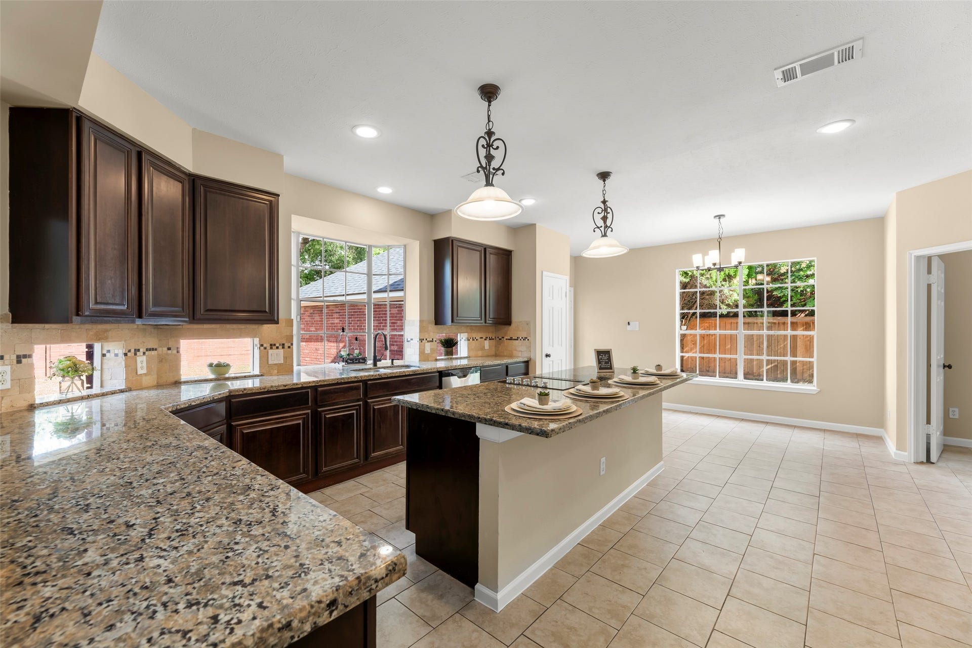 Granite countertops, dark wood cabinets, and a large island. It features pendant lighting, ample natural light from large windows, and tiled floors. Ideal for entertaining and family gatherings.