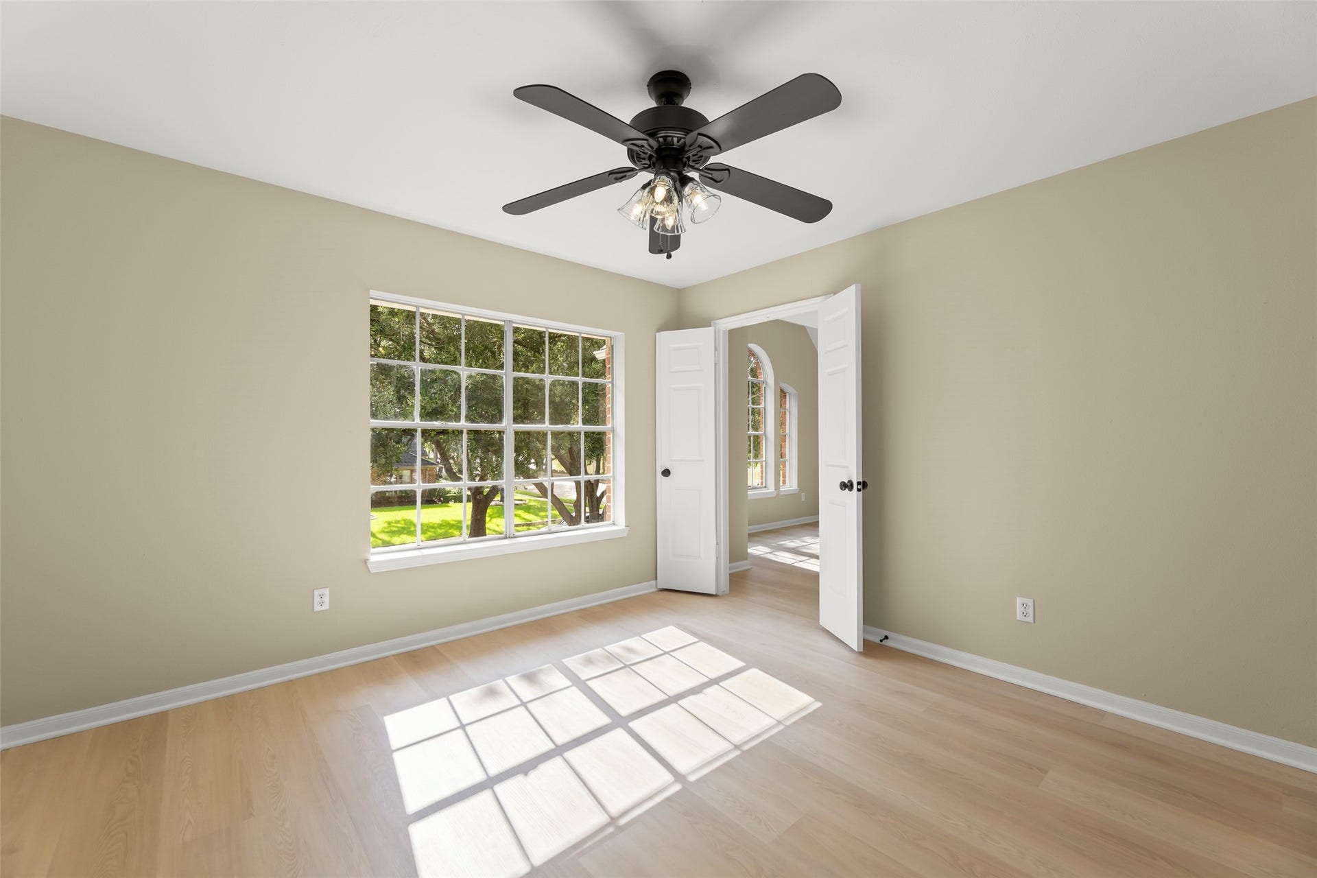 Bright room with large window, neutral walls, and light wood flooring. Features a ceiling fan and arched doorway, offering a view of lush greenery outside.