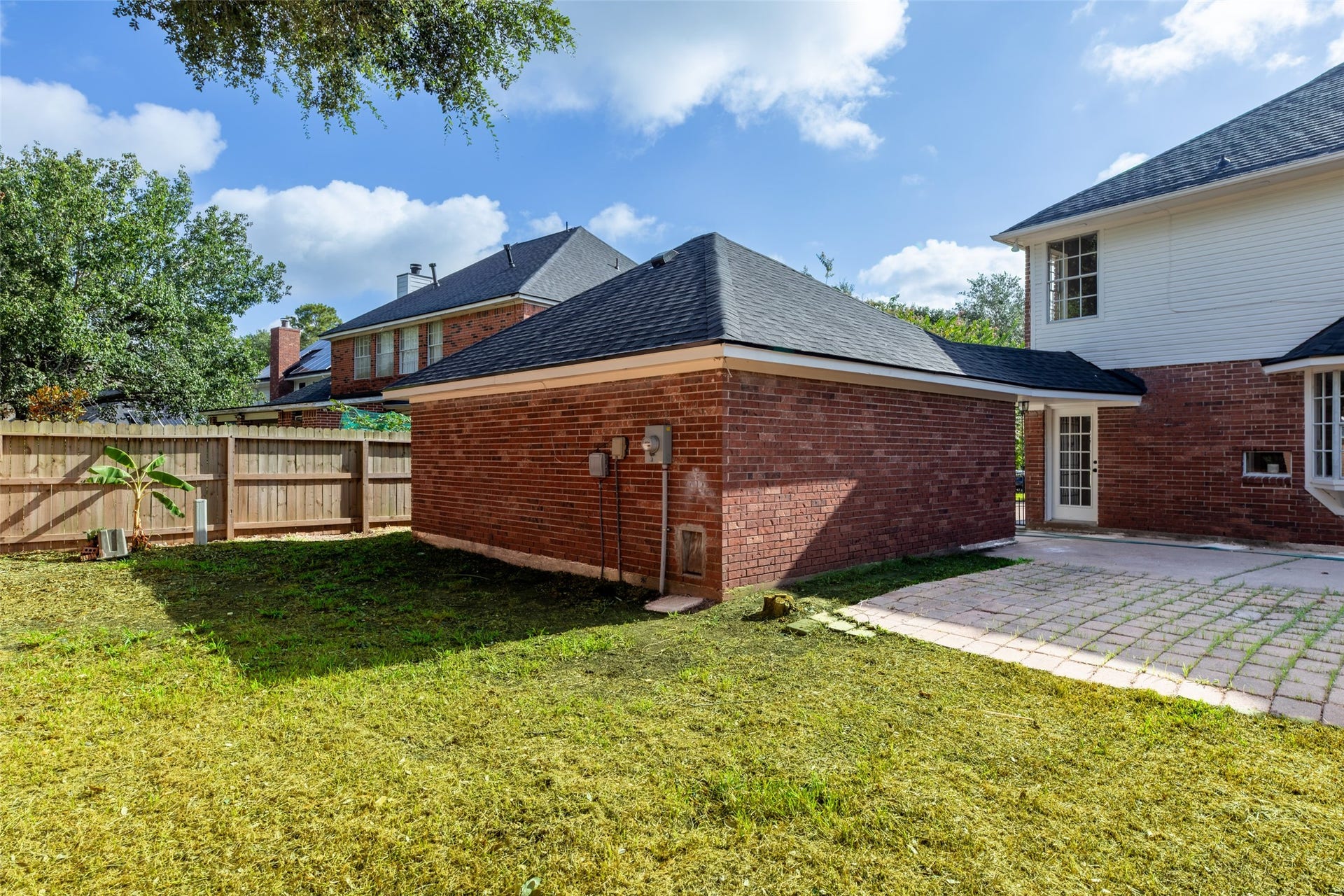 Well-maintained lawn and a small patio area. The house features red brick walls and a black roof, with a wooden fence providing privacy. There are a few trees and plants, adding greenery to the space.