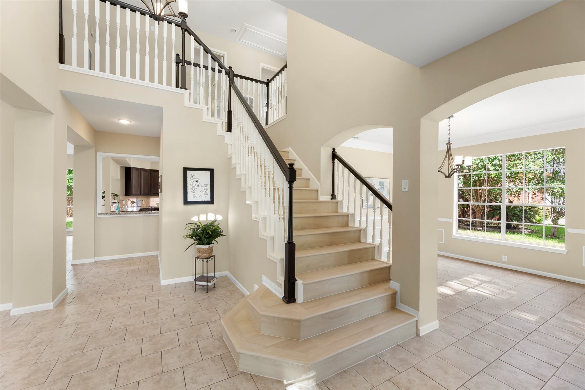 Open foyer with a stylish staircase, tiled floors, and ample natural light. Adjacent rooms offer versatile living spaces, and large windows overlook a lush garden.