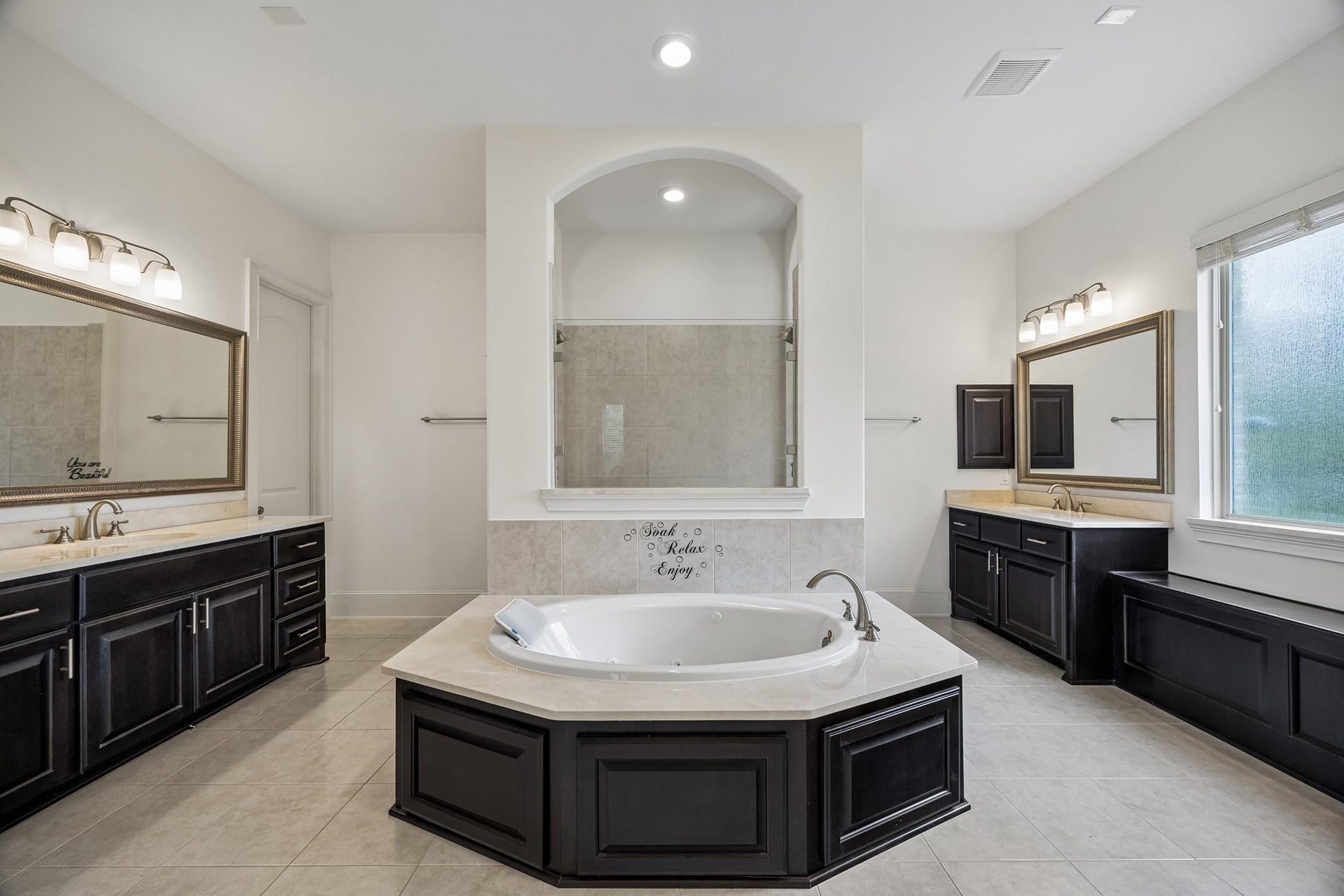 5018 Southampton Road, Missouri City, TX 77459 This primary bedroom bathroom features a central soaking tub, dual vanities with elegant dark cabinetry, a spacious walk-in shower, all enhanced by natural light and easy access to two walk in closets.