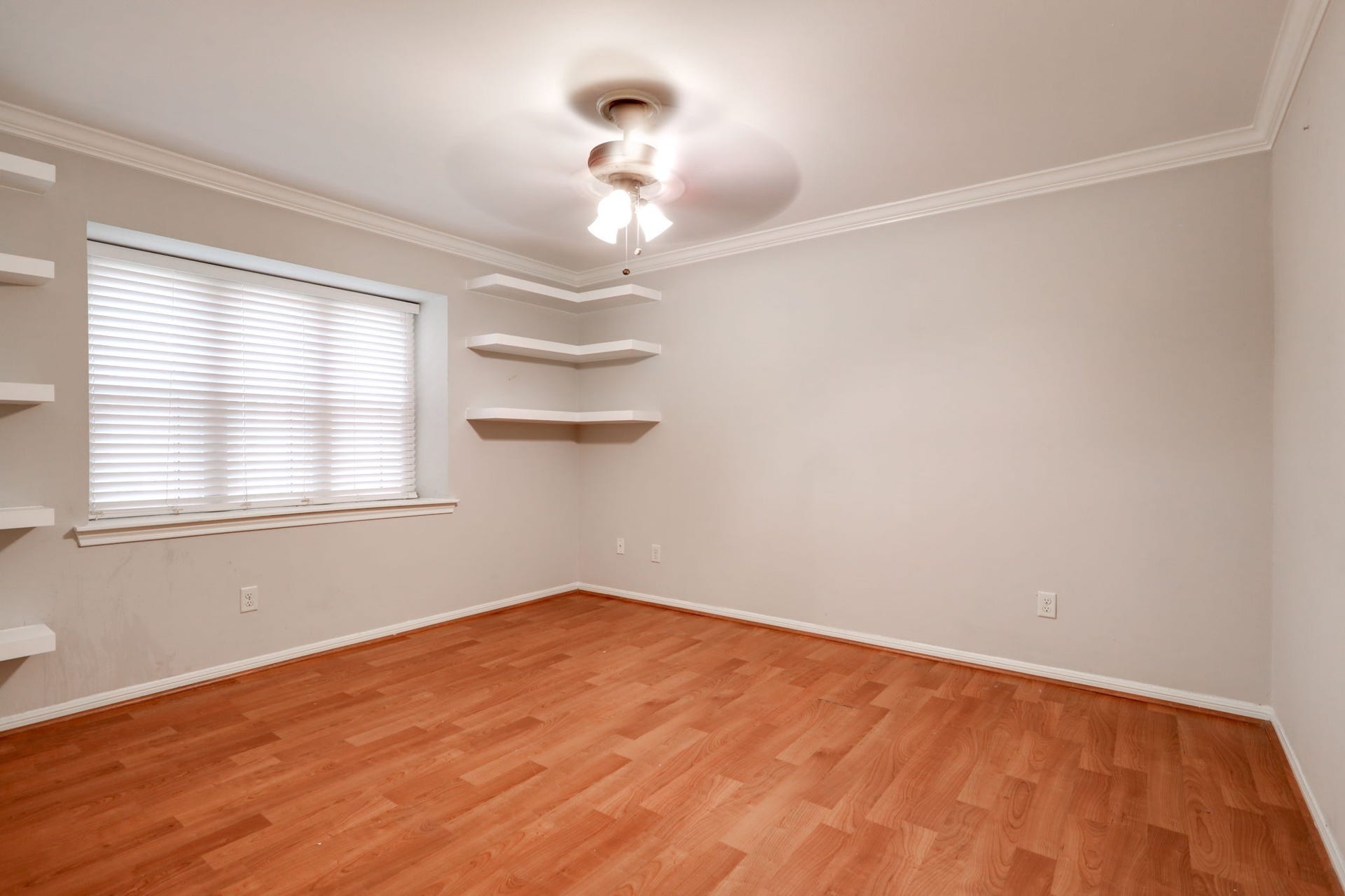 2912 Camelot Lane, Missouri City, TX 77459 Warm wood-look laminate flooring contrasts with neutral walls and white trim in the secondary bedroom. A wide window with blinds brings in natural light, while crown molding, a ceiling fan, and wall-mounted shelves add detail and function. The room offers flexibility for bedroom furnishings, a desk, or a reading corner.