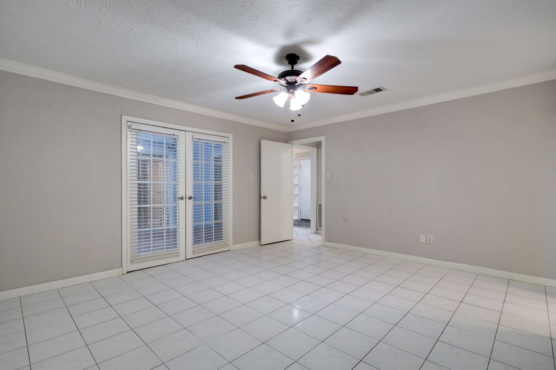2912 Camelot Lane, Missouri City, TX 77459 French doors with blinds frame a view into the central atrium, giving the primary bedroom a distinctive connection to the heart of the home. Tile flooring, crown molding, and a ceiling fan complement the neutral interior. An adjoining en-suite bathroom adds convenience to this inviting bedroom retreat.