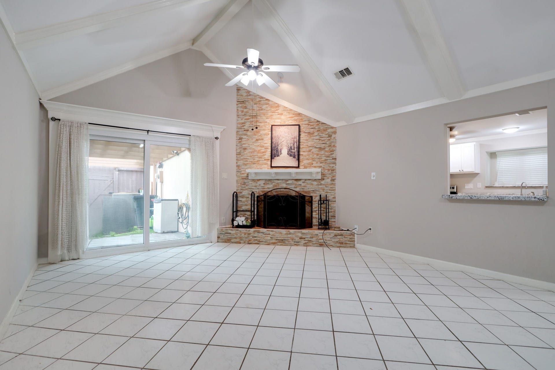 2912 Camelot Lane, Missouri City, TX 77459 Vaulted ceilings and exposed beams give this spacious living room an airy, expansive feel. The floor-to-ceiling stone fireplace surround and raised hearth create a striking focal point, while sliding glass doors open to the private fenced patio. Tile flooring, ceiling fans, and a kitchen pass-through combine architectural character with everyday practicality.