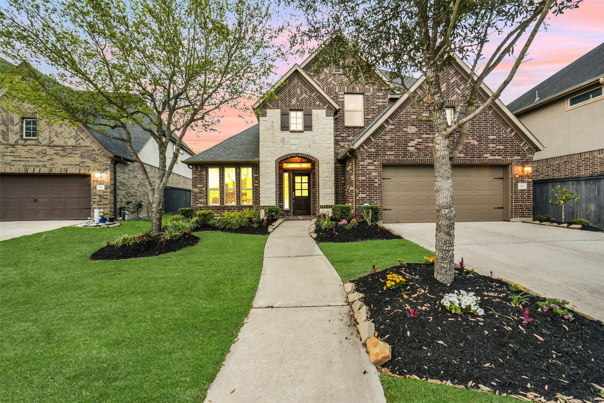 Charming two-story brick home with a stone accent facade, featuring a two-car garage and a beautifully landscaped front yard. The pathway leads to a welcoming entrance, highlighted by large windows and mature trees.