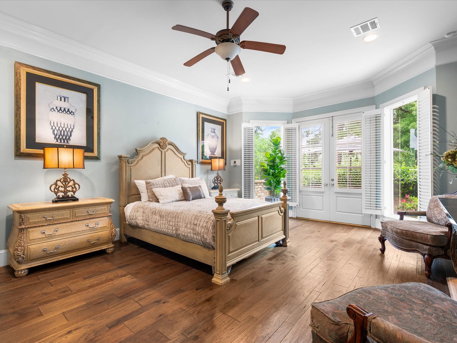 Bathed in natural light, the expansive primary suite offers soaring ceilings, crown molding, gleaming wood floors, and a comfortable sitting area. French doors open to the gardens beyond, creating a private sanctuary designed for rest and relaxation.