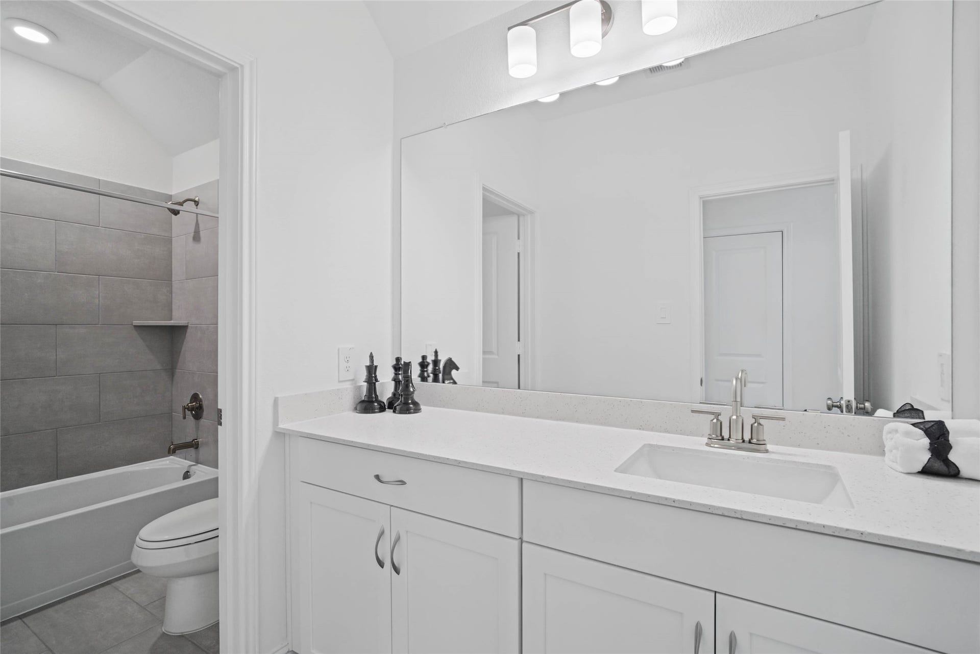 Secondary bathroom featuring an extended vanity cabinet with single bowl sink, brushed nickel hardware, mirror and vanity lighting, tub/shower combination with tile surround and tile flooring.