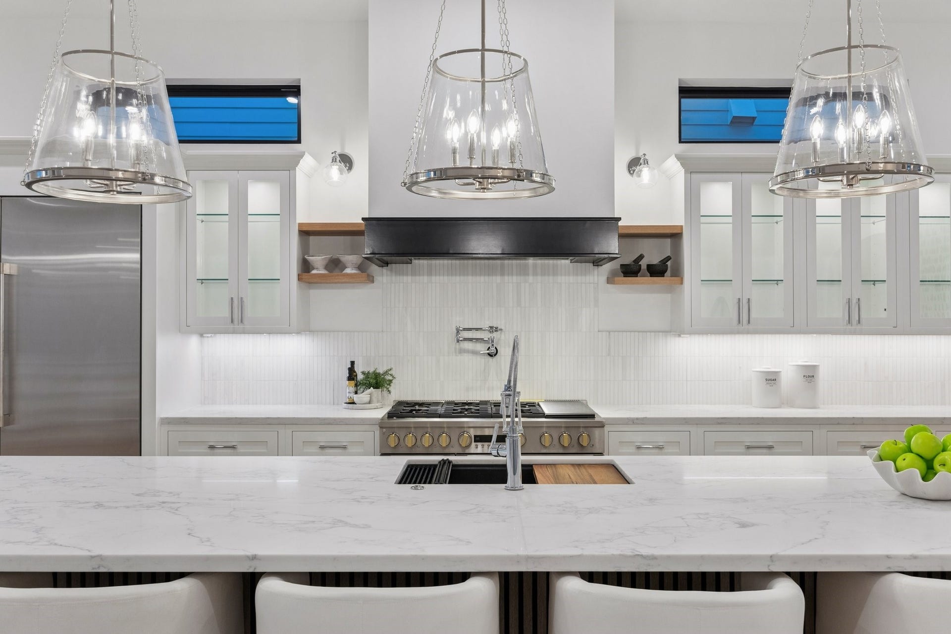 A closer view of this gorgeous kitchen showcases Slab Quartz Counter, 1 x 6 Honed Stacked Marble Backsplash, 33 Inch Undermount Single Basin Sink in Quartz Composite Finish, Pull-Down Faucet in Polished Chrome and Top Switch Disposal, 24" Pot Filler in a Polished Chrome Finish, White Oak Floating Shelves with 6" LED Sconces  in a Chrome Finish with Clear Shades, LED Under-Cabinet Lighting and Under-Cabinet PlugMold. Inset Cabinetry with Glass Fronted Doors and Shelves with Lighting on the Upper Cabinets.