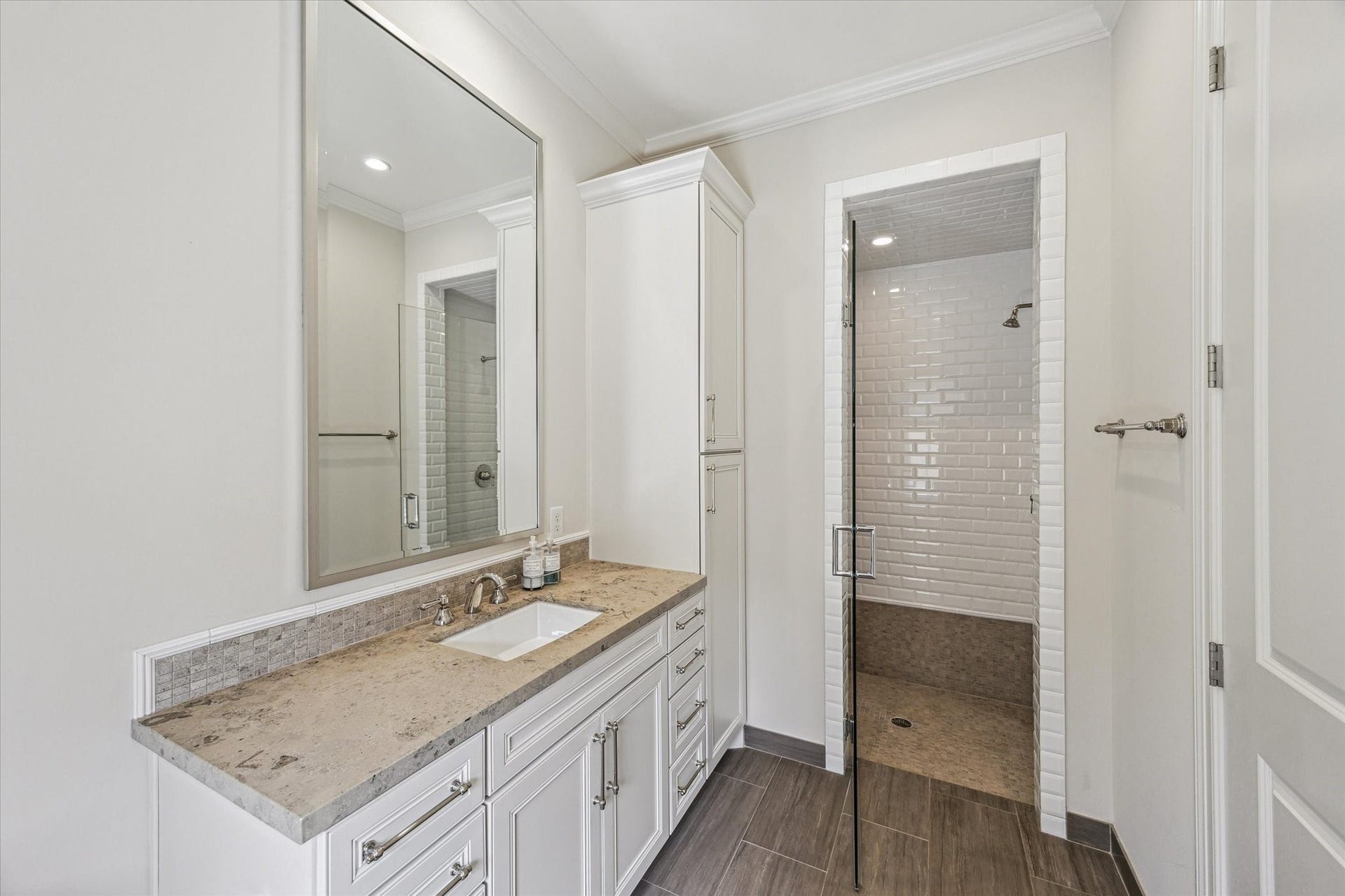2413 Mimosa Drive, Houston, TX 77019 The en-suite bath keeps the palette quiet and timeless with stone counters, white cabinetry, polished hardware, and a glass-enclosed shower lined in classic white tile. A tall cabinet adds useful storage without taking away from the rooms streamlined feel.