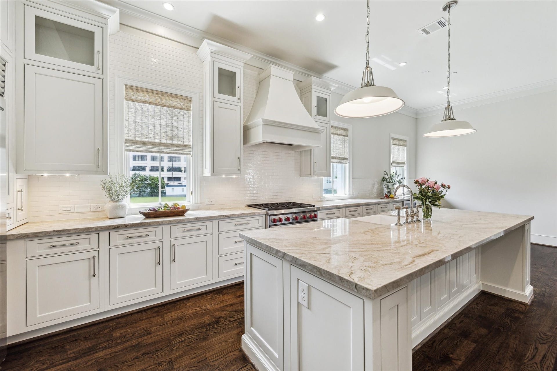 2413 Mimosa Drive, Houston, TX 77019 There is a quiet symmetry to the kitchen: tall custom cabinetry, glass-front display cabinets, woven shades, and a sculptural vent hood are arranged around the professional-style range. Stone counters and a full-height tile backsplash keep the palette light against the dark floors.
