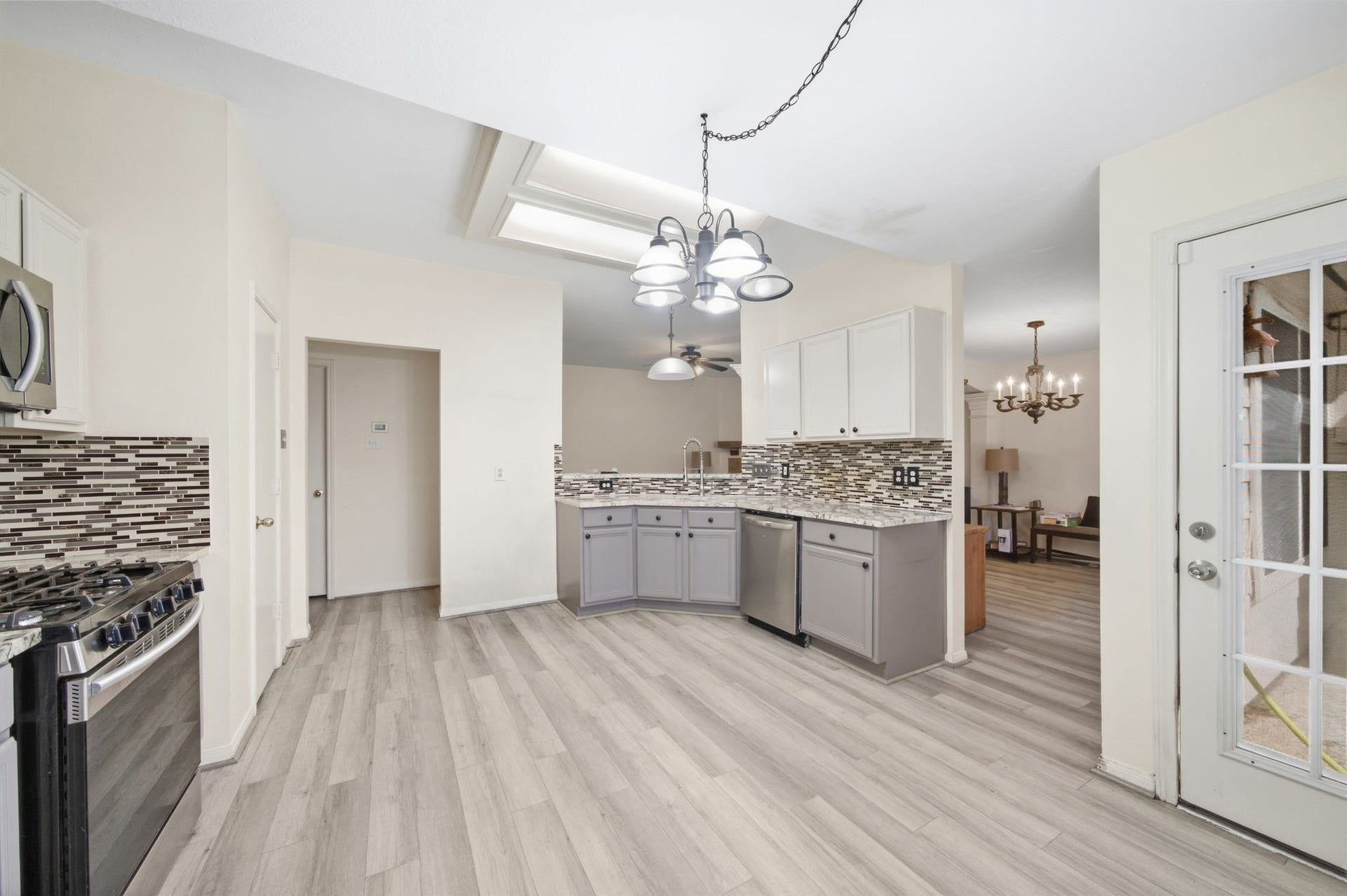 A tiered chandelier illuminates this spacious breakfast area and kitchen hub, complemented by low-maintenance wood-look floors. Two-tone cabinetry and granite countertops frame a stainless steel dishwasher and corner sink with sightlines into the living space, while a multi-pane door provides direct access outside.