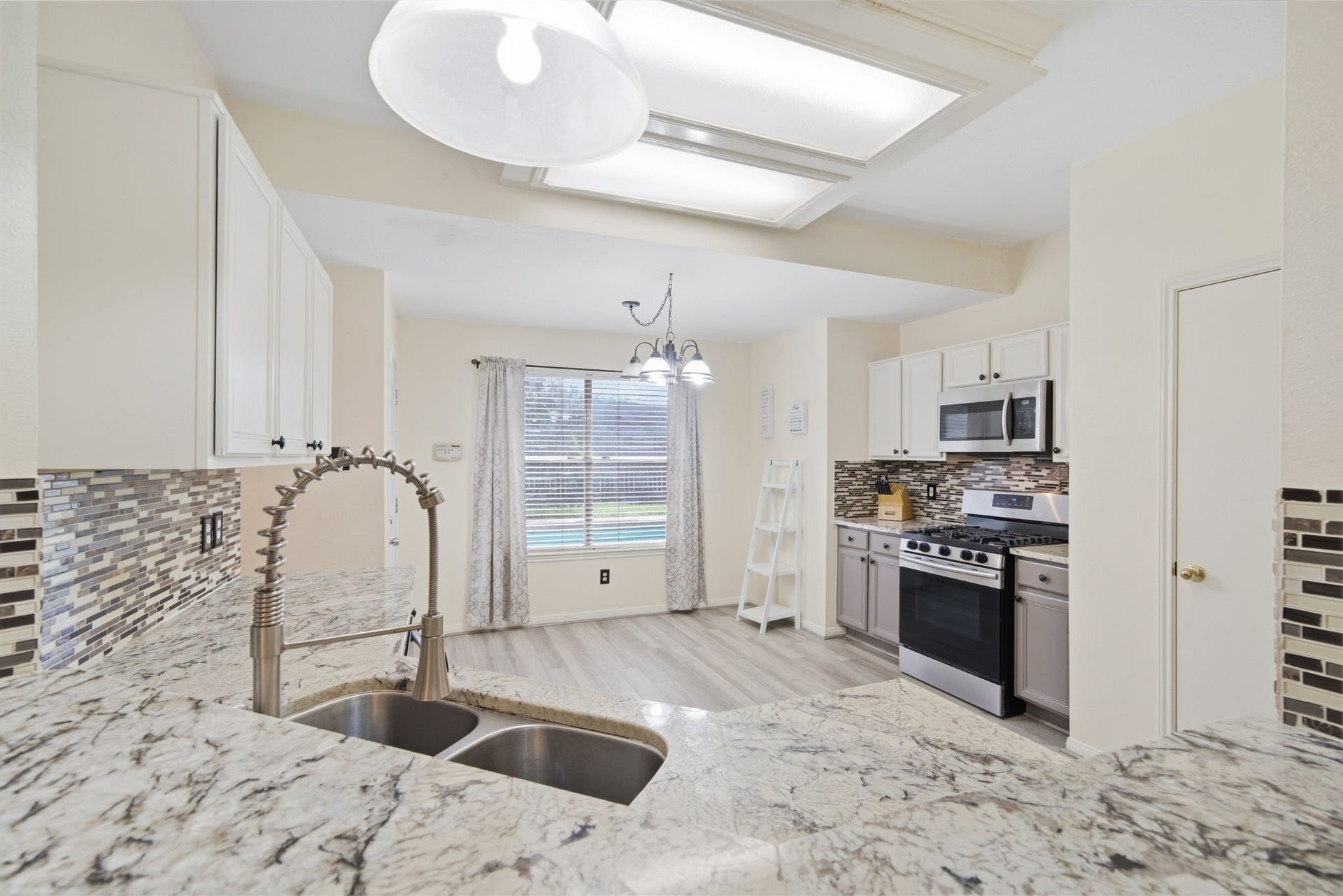 Granite countertops and an undermount dual sink create a functional workspace, with an open peninsula overlooking the breakfast area. A wide window brings natural light across low-maintenance flooring, while sleek appliances and a nearby pantry door add everyday convenience.