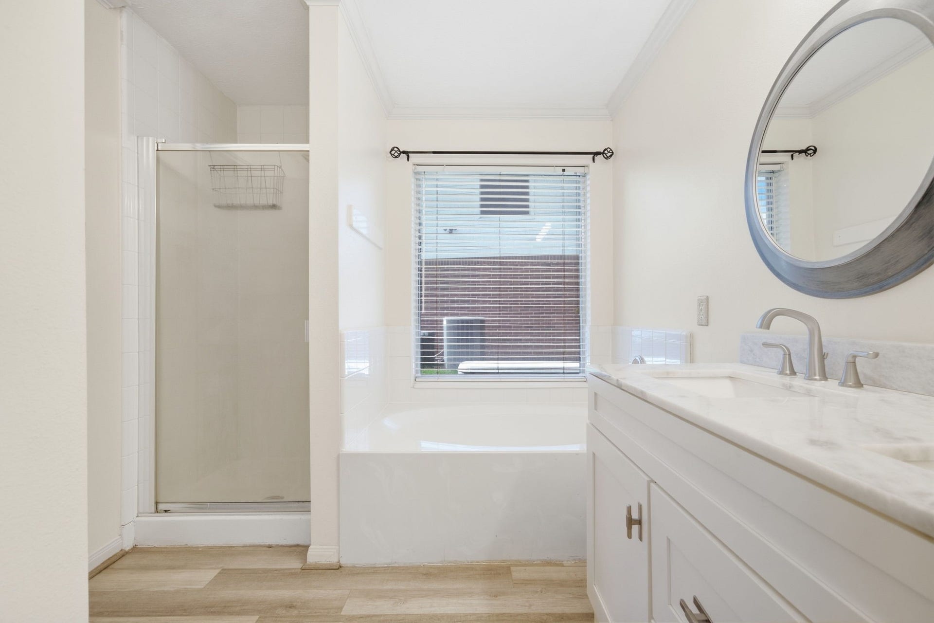 Crisp white surfaces and warm wood-look floors define this bright primary ensuite bath. A tiled soaking tub rests beneath a sunny picture window, positioned alongside a glass-door walk-in shower and an updated marble-look vanity with brushed nickel hardware.