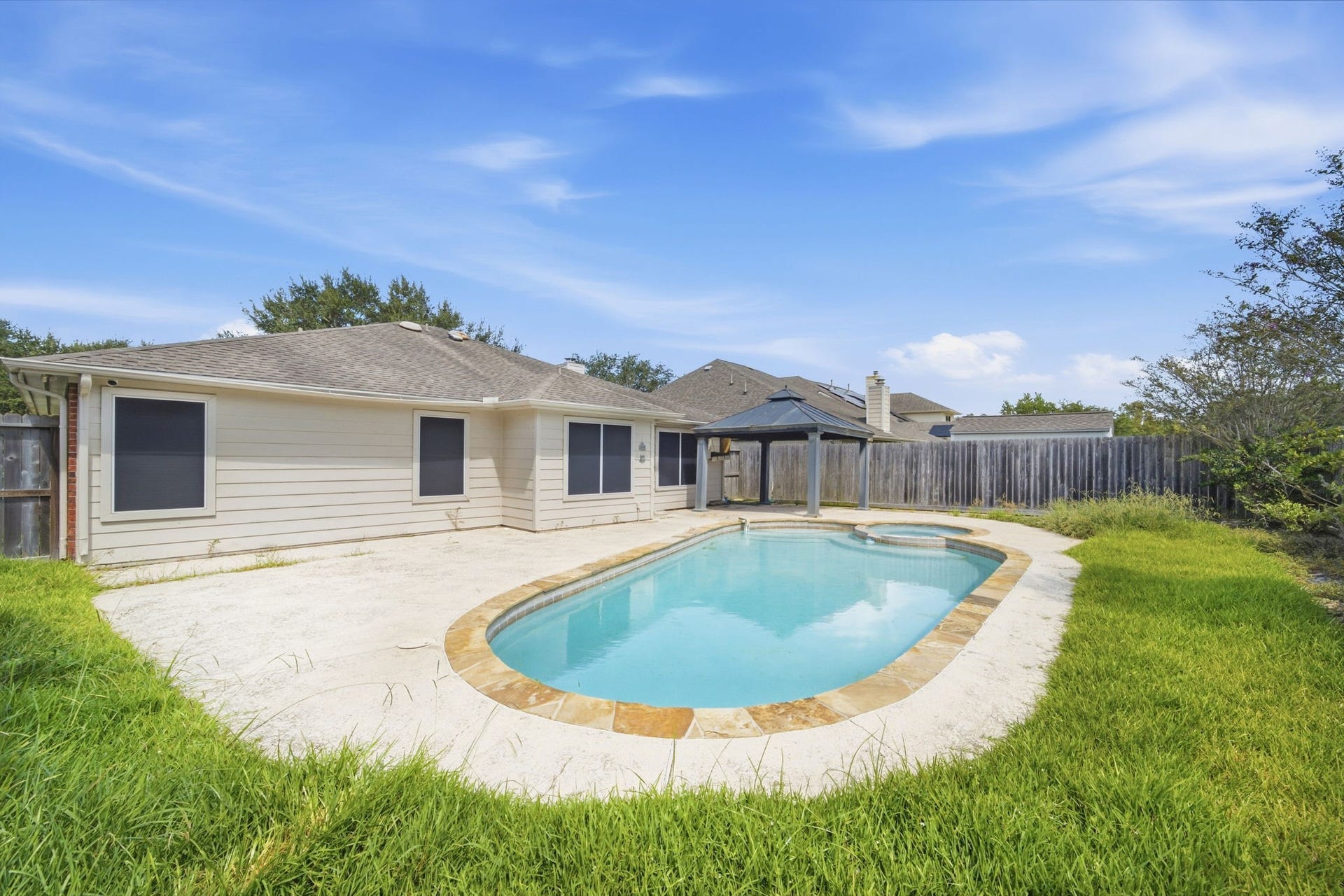 Stone coping frames the sparkling in-ground pool and attached hot tub, creating an inviting backyard retreat primed for outdoor leisure. A shaded gazebo canopy and generous concrete decking provide practical space for lounge seating, while perimeter privacy fencing encloses the level lawn.