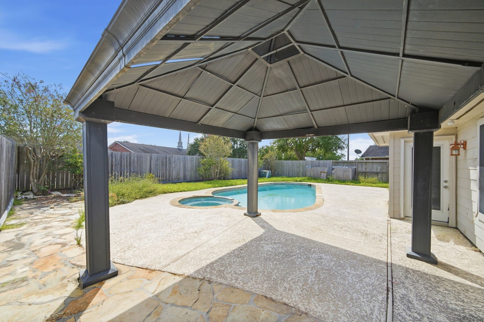 Flagstone accents meet expansive textured concrete decking beneath a sturdy shaded pavilion, delivering an ideal outdoor venue for open-air dining. Beyond the shelter, an in-ground swimming pool and adjoining hot tub sit framed by a wood privacy fence, establishing solid value and relaxed outdoor living.