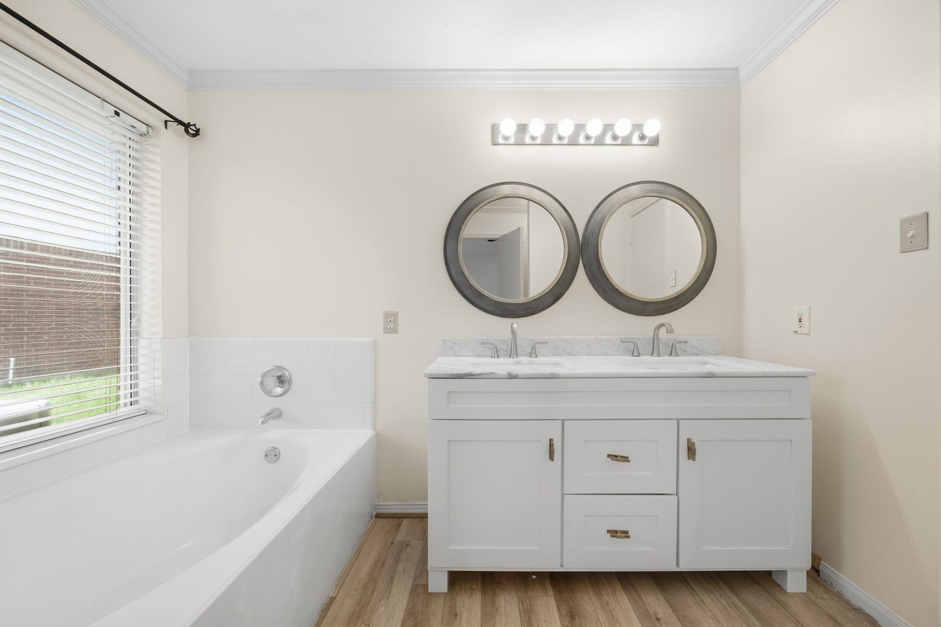 Relax in the private primary ensuite bath, complete with a deep soaking tub positioned beside a sunny window. A crisp white vanity features dual undermount sinks, marble-patterned countertops, dual circular mirrors, and low-maintenance wood-look floors, providing optimal functionality for daily routines.
