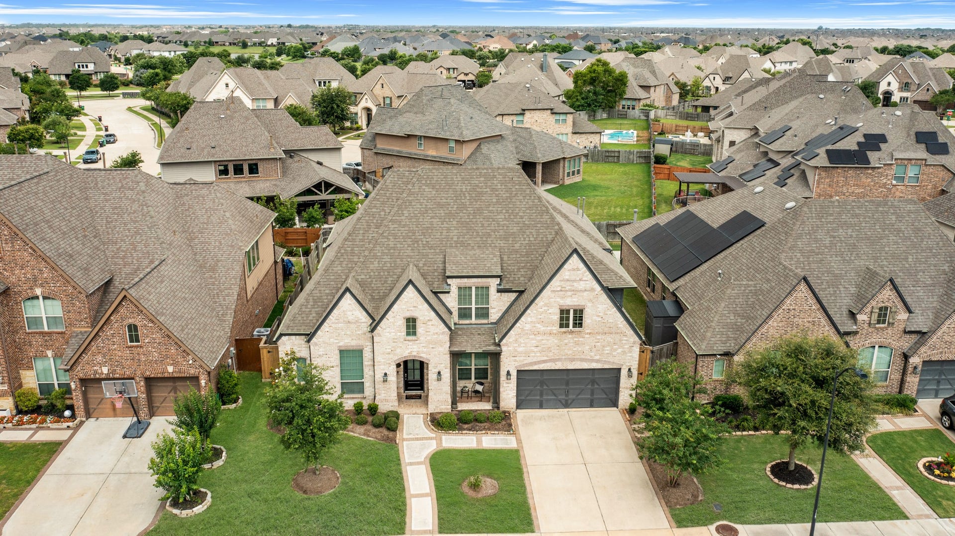 An elevated front-facing drone view captures the home's striking architectural peaks, extensive roofline, and prime location nestled within the beautifully manicured Elyson neighborhood.