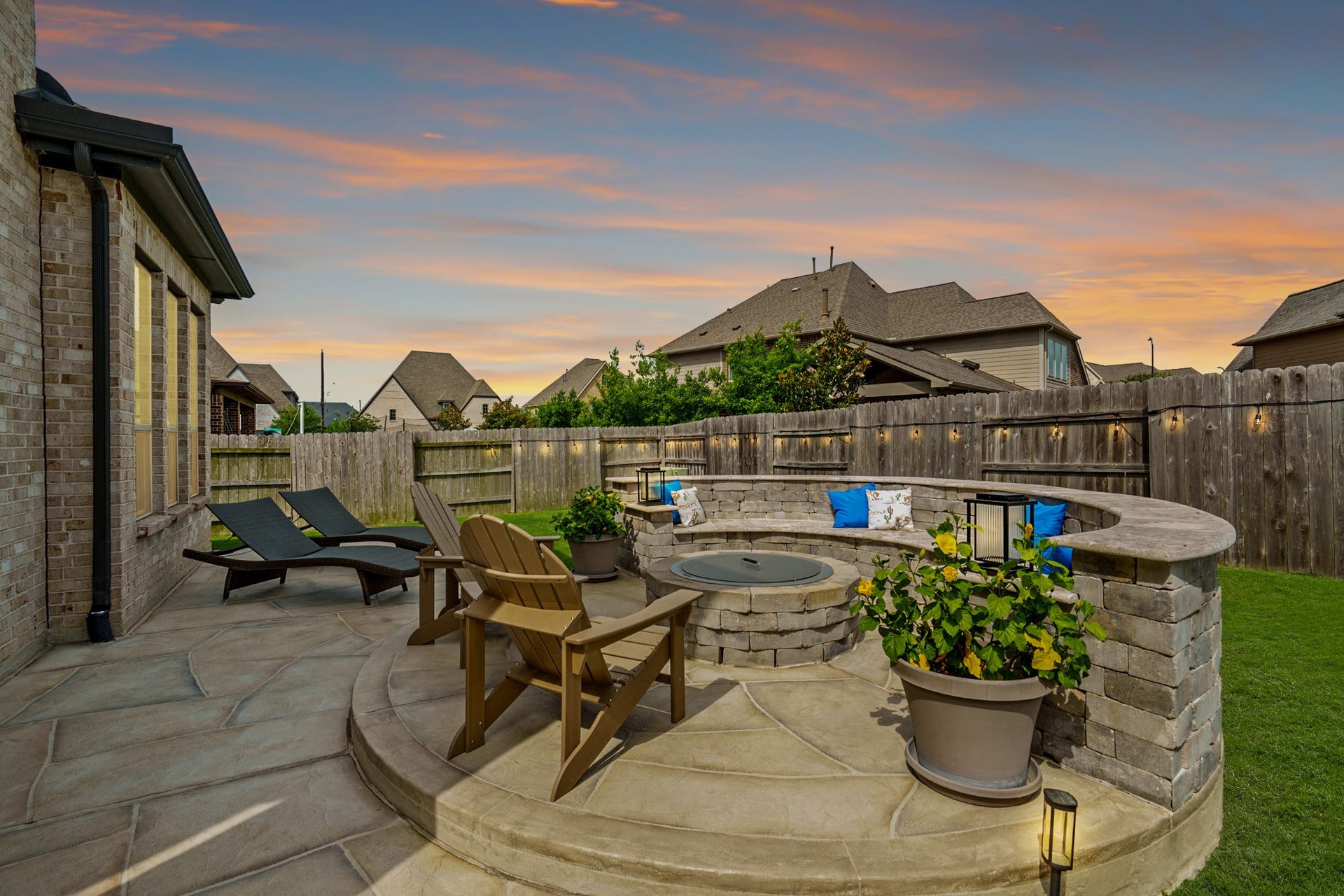 A spectacular dusk view of the custom stone fire pit area, featuring integrated curved bench seating, elegant flagstone paving, and ambient string lighting along the fence line.