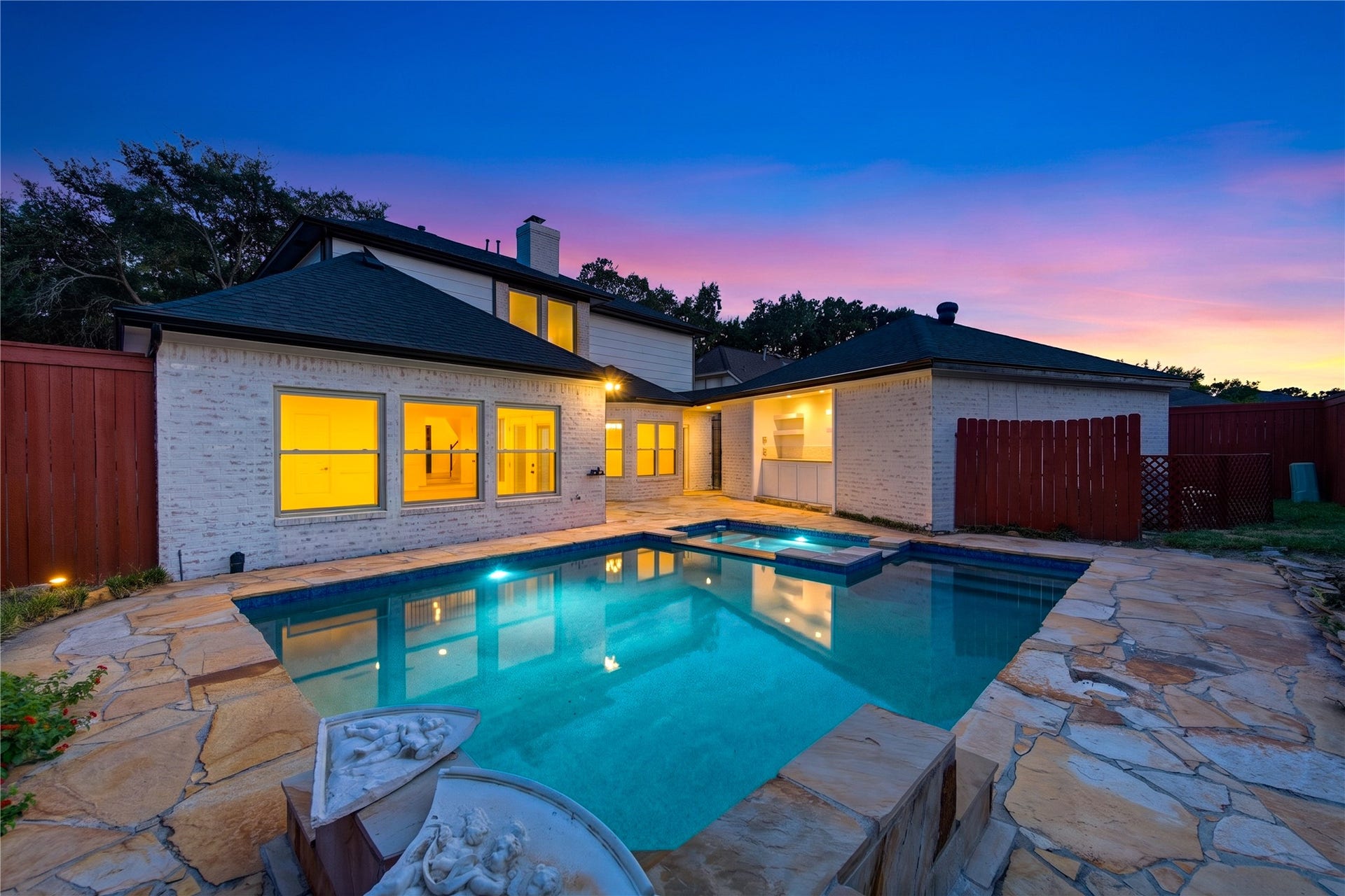 Another twilight view of the backyard showcases the sparkling pool and generous surrounding patio, creating an inviting extension of the home for sunset swims, weekend barbecues and evenings spent relaxing outdoors with family and friends.
