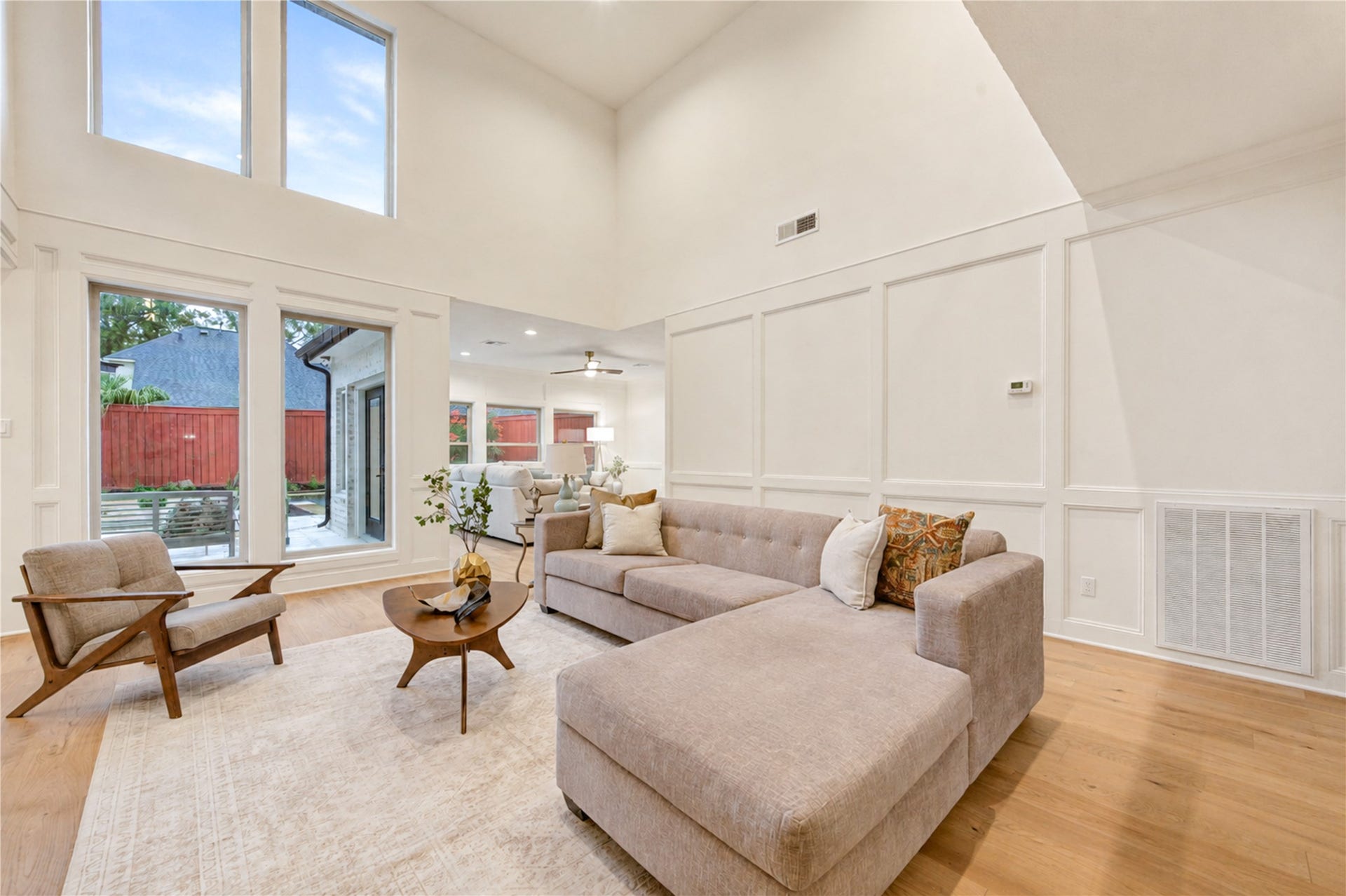 Formal living room showcases dramatic two-story ceilings, French Oak floors and elegant wood-paneled walls that add architectural detail, while expansive windows fill the space with natural light and offer views toward the backyard.