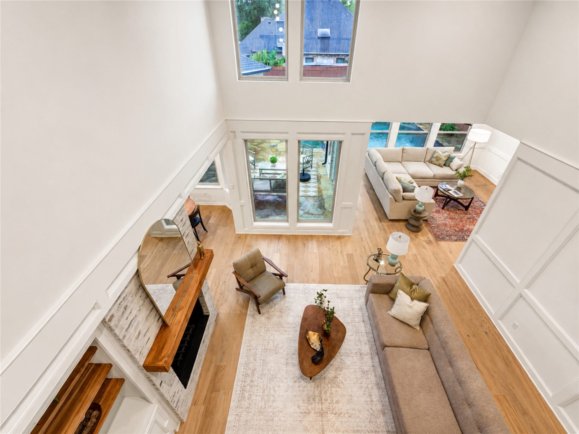 Dramatic two-story living room featuring soaring ceilings, abundant natural light, French Oak floors and a striking fireplace, with expansive windows overlooking the backyard and pool.