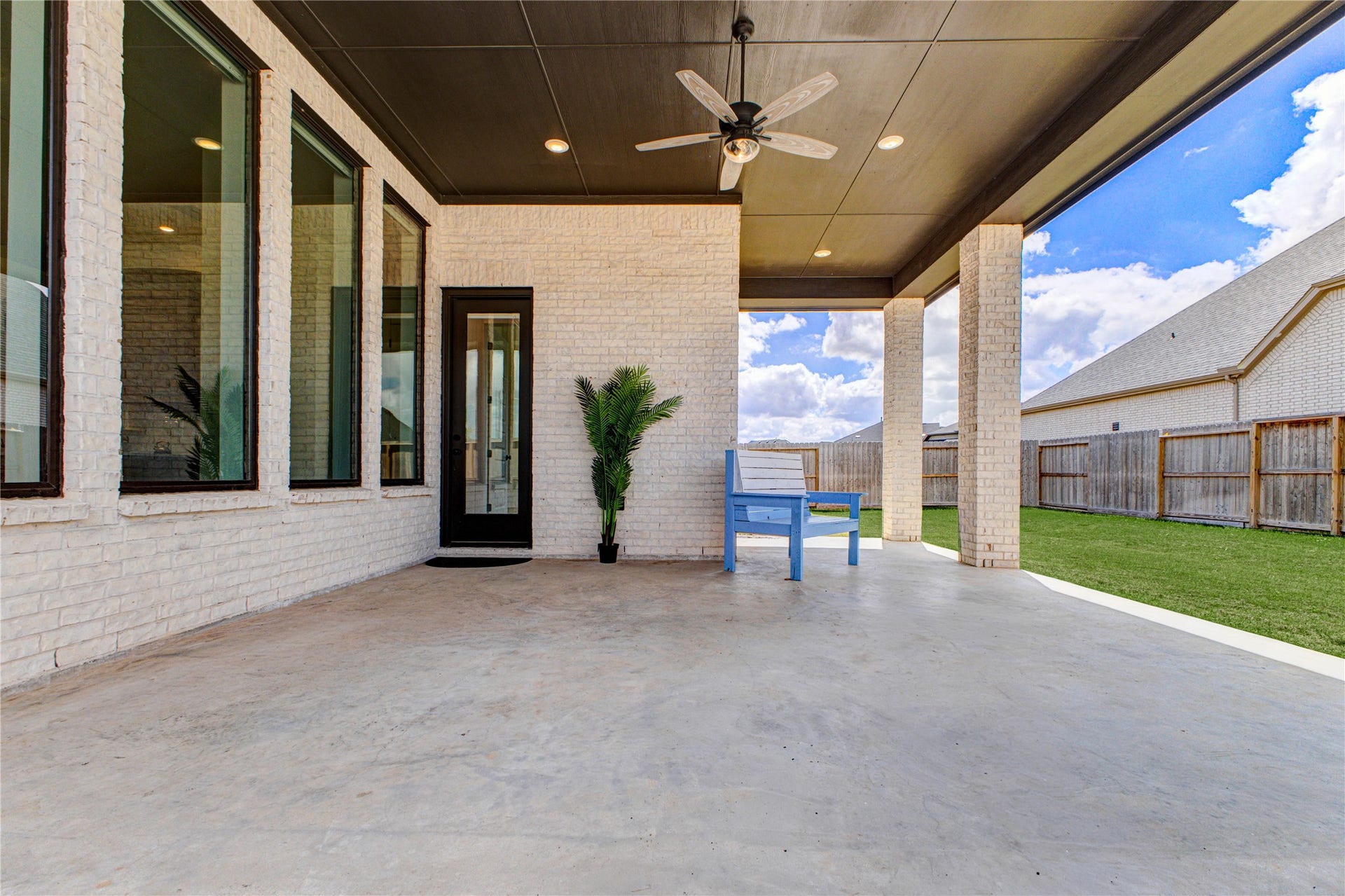 4510 Windmill Landing Lane, Fulshear, TX 77441 Spacious covered patio with a ceiling fan, large windows, and a door leading inside. The area features a simple potted plant and a blue bench, overlooking a well-kept lawn, perfect for outdoor relaxation.