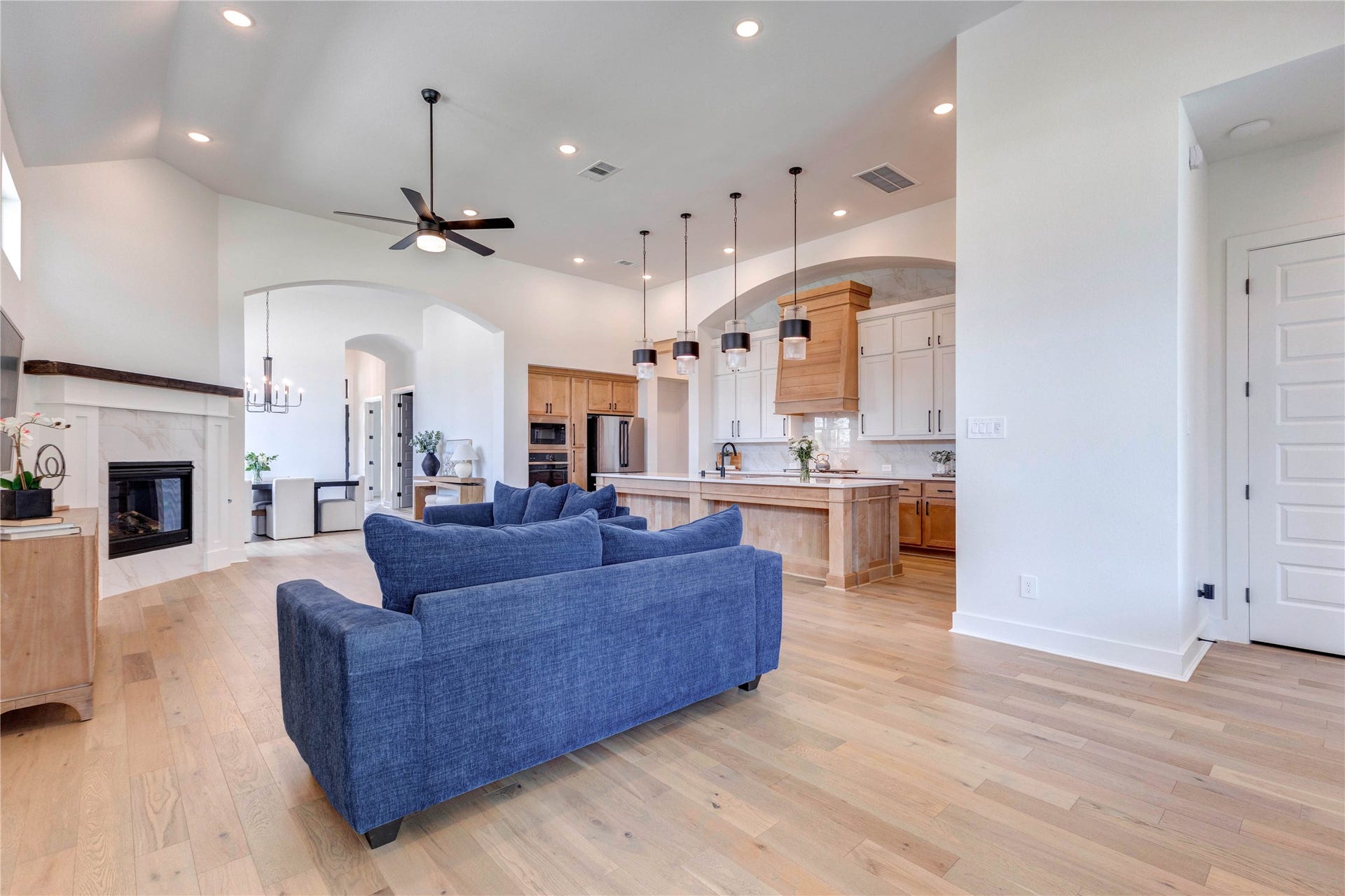 4510 Windmill Landing Lane, Fulshear, TX 77441 This spacious living room features high ceilings, recessed lighting, and a ceiling fan. It includes a cozy fireplace, large windows for natural light, and stylish blue seating. The open layout leads to a dining area with a modern chandelier, enhancing the home's airy feel.