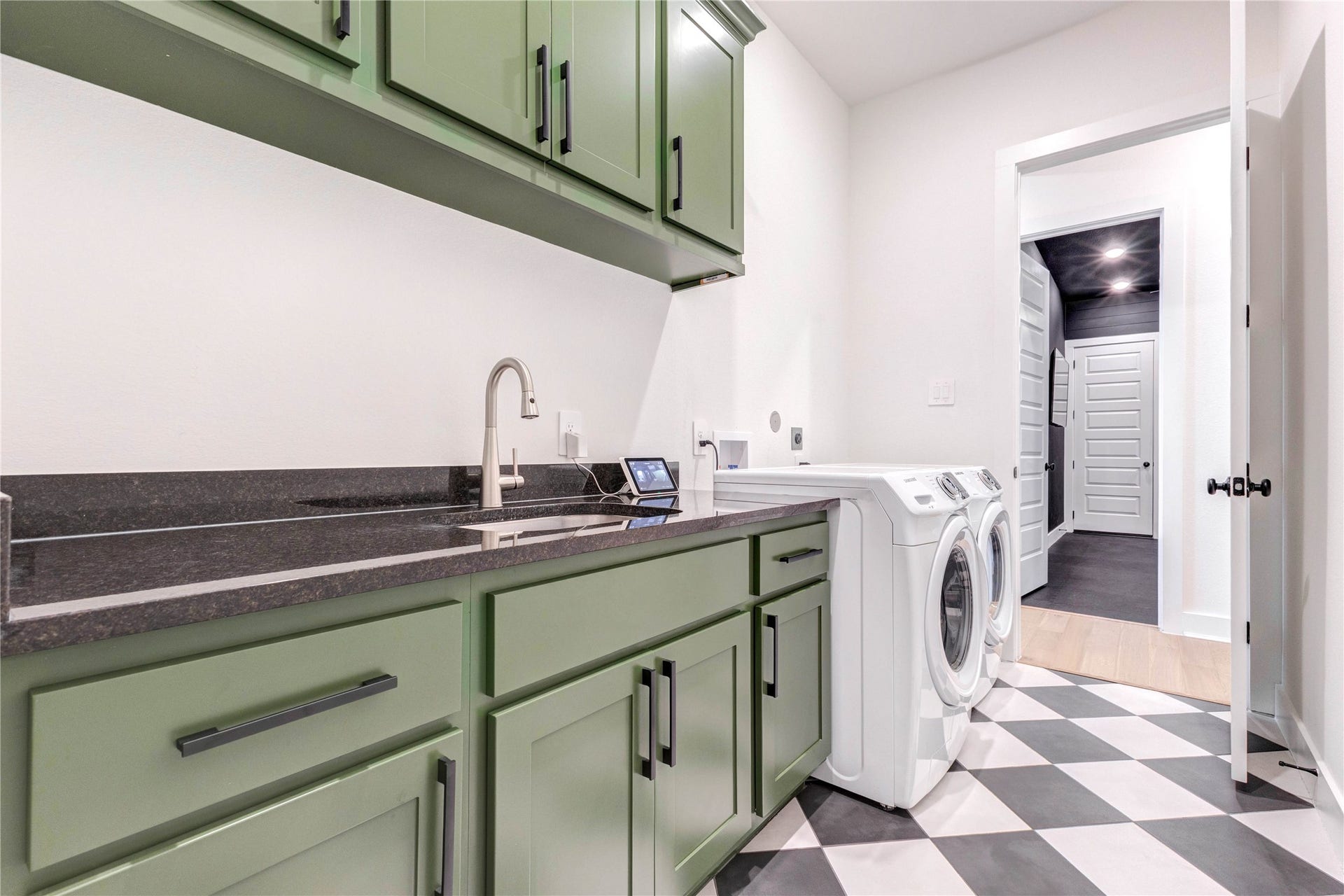 4510 Windmill Landing Lane, Fulshear, TX 77441 This laundry room features stylish green cabinetry, a sleek black countertop, and modern appliances. The space is well-lit and has a checkered floor, offering a functional and attractive area for laundry tasks.