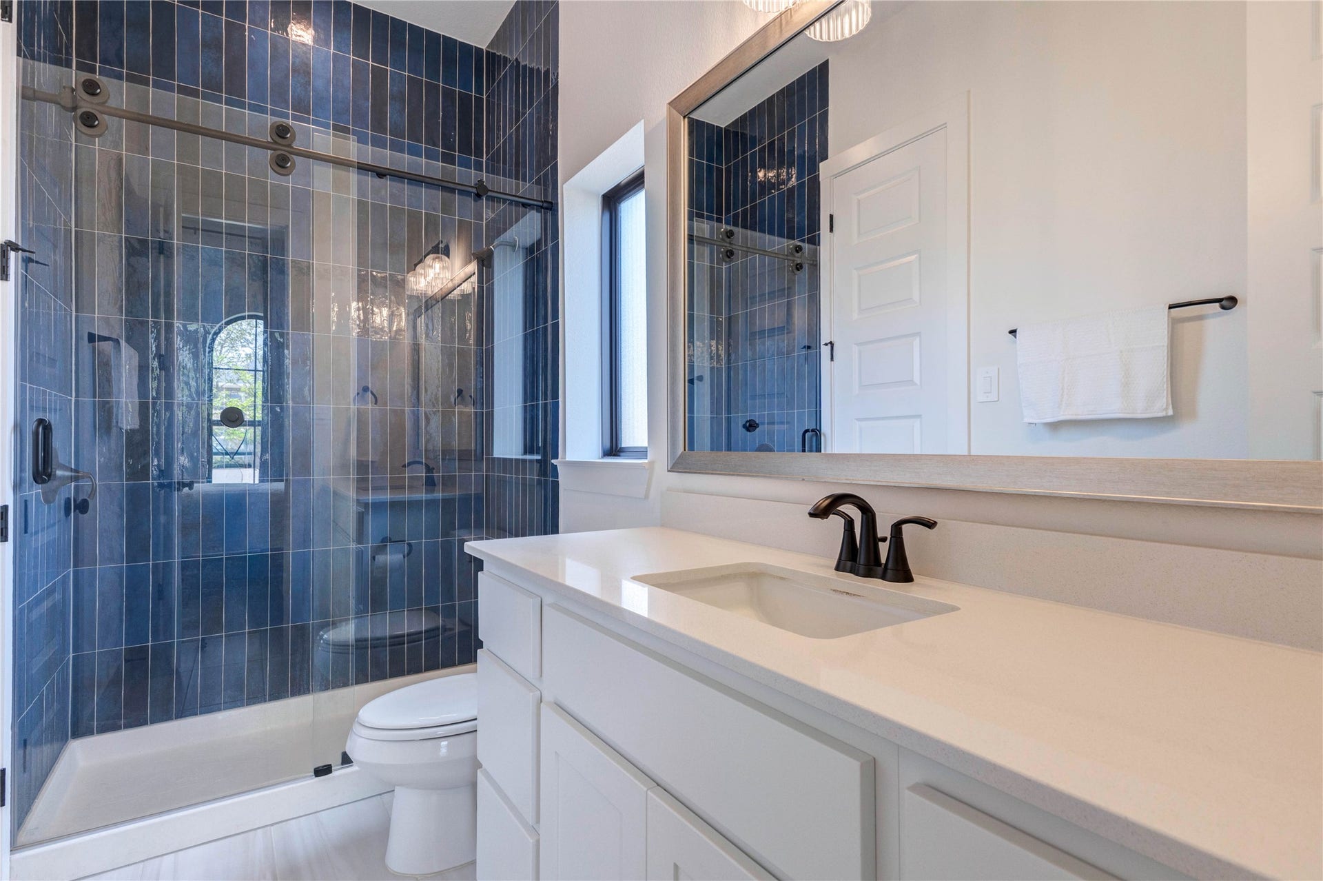 4510 Windmill Landing Lane, Fulshear, TX 77441 This bathroom features a sleek, modern design with a double vanity in deep blue tile, black fixtures, and a large mirror. The space is bright with white walls and wood flooring, offering a clean, stylish look.