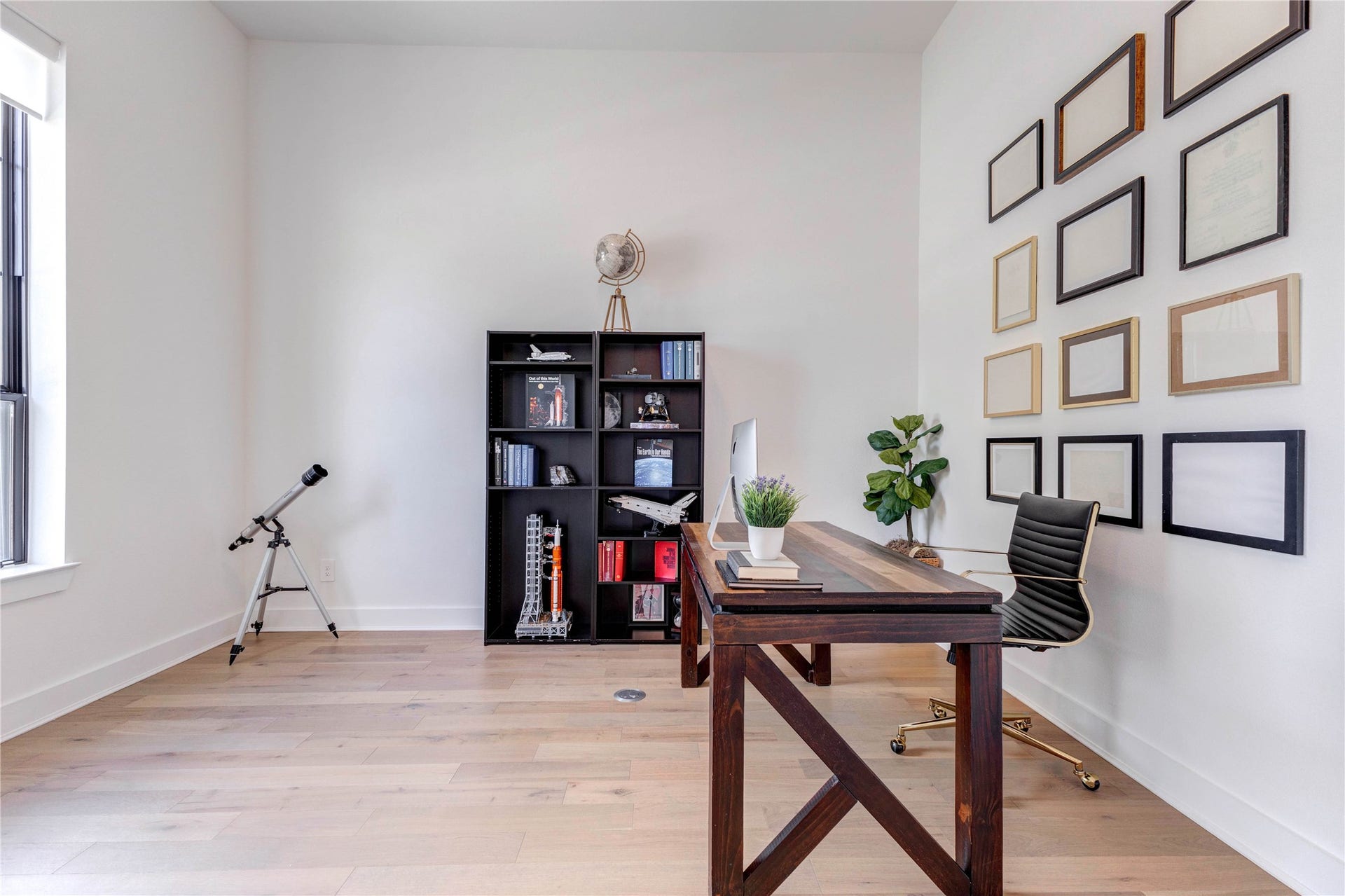 4510 Windmill Landing Lane, Fulshear, TX 77441 Modern home office with a sleek wooden desk, stylish chair, and a black bookshelf. Decor includes a telescope by the window, a plant, and a wall display of empty picture frames. The room features light wood flooring and ample natural light.