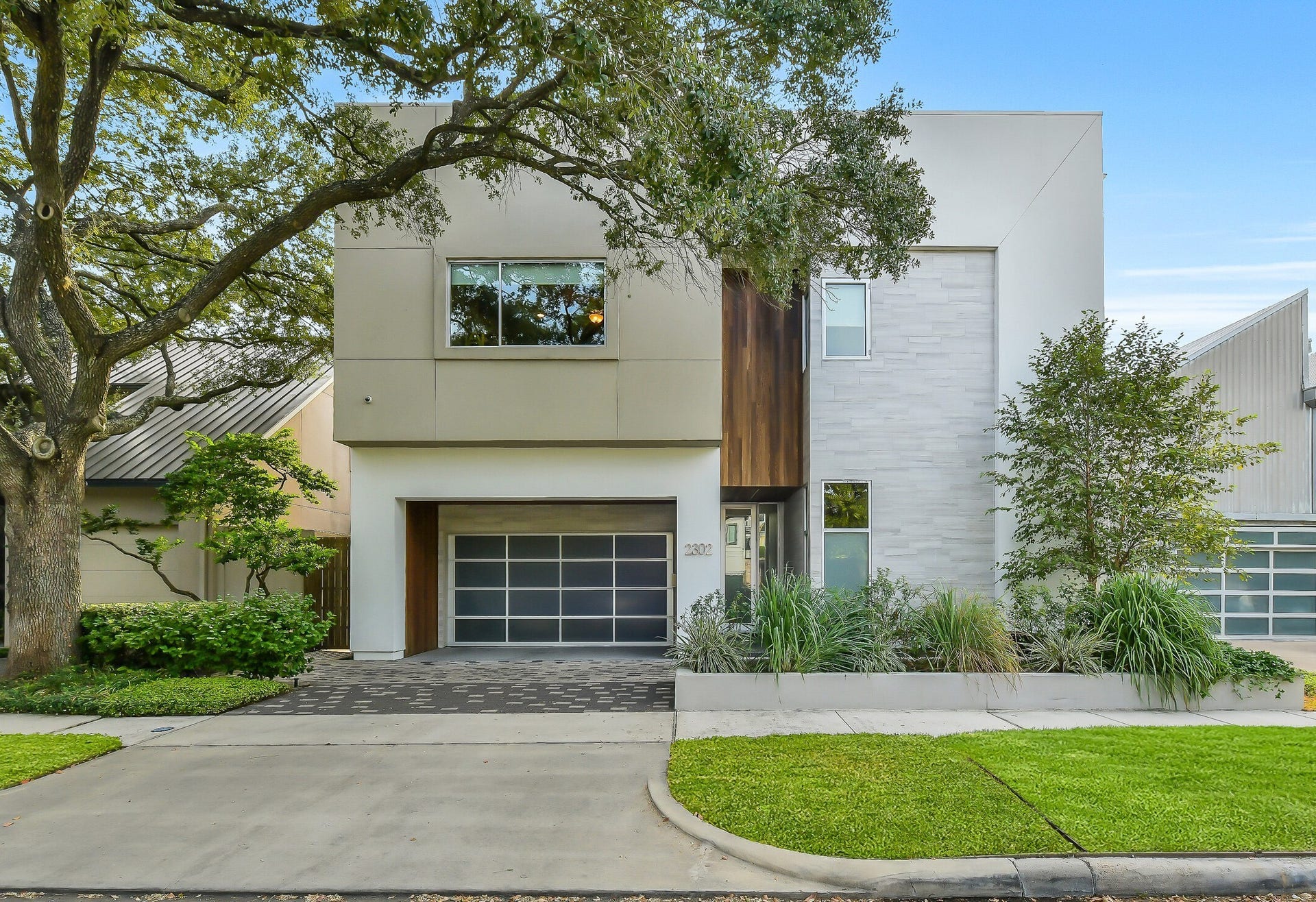 2302 Persa Street, Houston, TX 77019 This striking modern home captures immediate attention with its bold geometric facade, seamlessly blending sleek stucco, porcelain accents, and warm wooden paneling. Framed by a classic mature oak tree, the exterior creates a captivating balance between contemporary architecture and natural warmth.