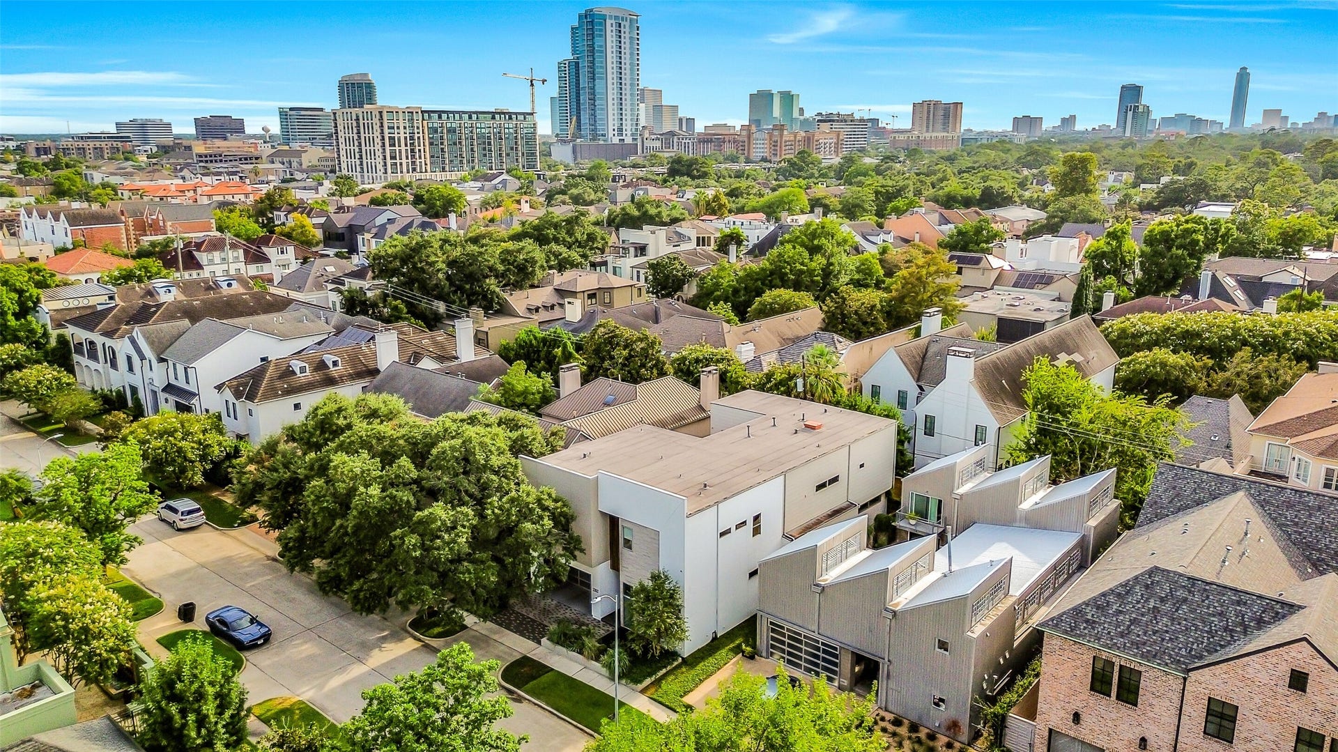 2302 Persa Street, Houston, TX 77019 This aerial view highlights the striking modern two-story residence nestled among established homes, mature trees, and nearby urban high-rises. The view emphasizes the property's unique contemporary design, private side courtyard, and prime locatin within a vibrant city neighborhod.