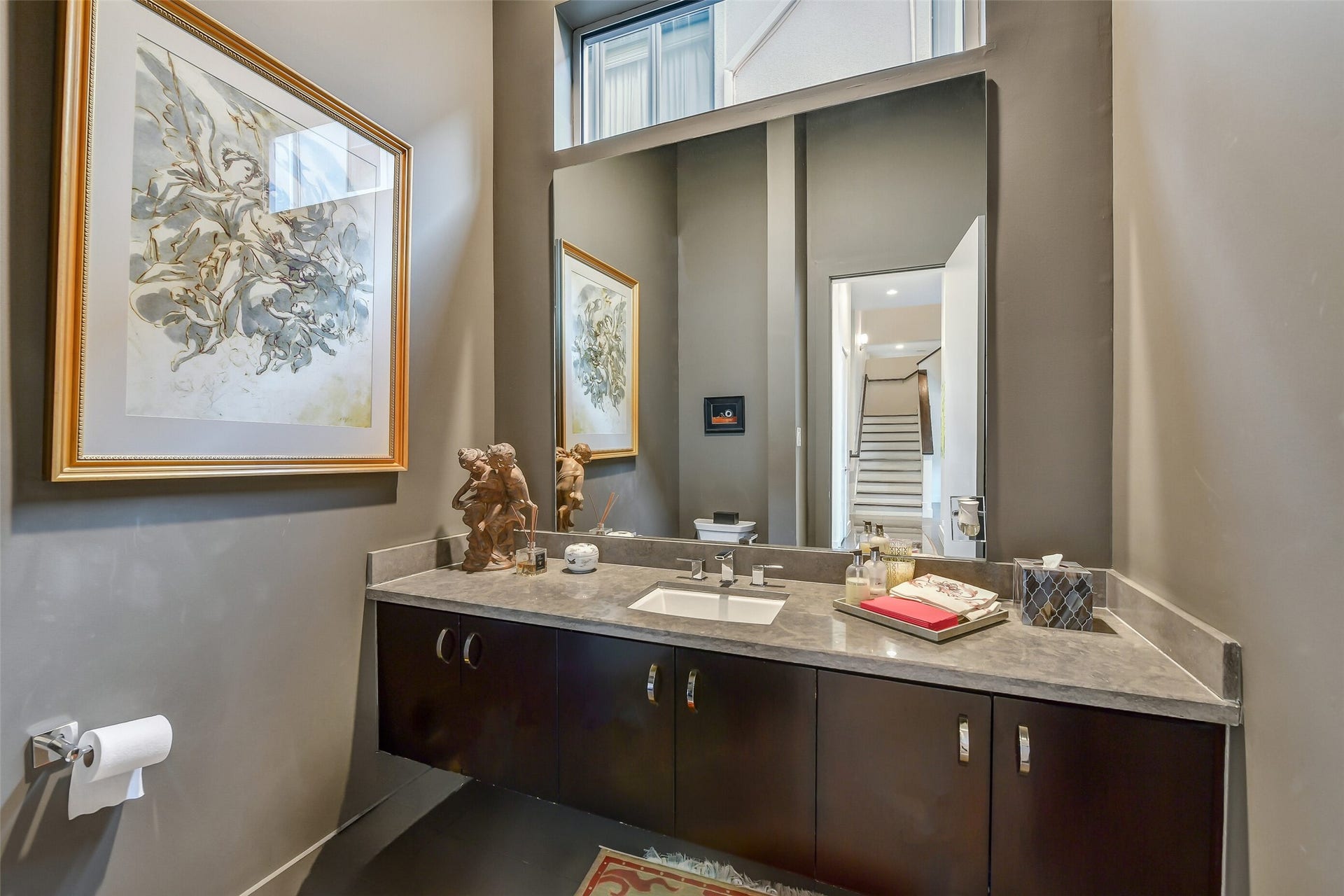 2302 Persa Street, Houston, TX 77019 This half bathroom features a floating dark-wood vanity topped with a stone countertop, a sleek undermount sink, and modern chrome fixtures. A tall, frameless mirror accentuates the room's height, while warm gray walls create a refined, contemporary powder room layout.