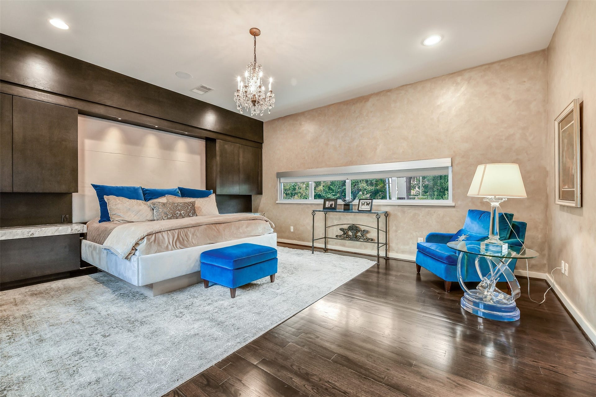 2302 Persa Street, Houston, TX 77019 The luxurious primary retreat pairs soft neutral tones with rich dark hardwoods and elegant modern lighting to create a serene sanctuary. A sophisticated dark-wood built-in accent wall frames the bed, featuring integrated overhead lighting and sleek cabinetry.