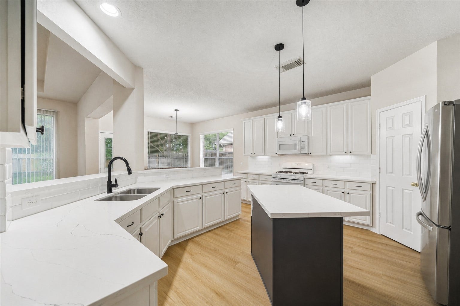 SPACIOUS KITCHEN WITH CENTER ISLAND, WHITE COUNTERTOPS, SUBWAY TILE BACKSPLASH, AND PENDANT LIGHTING OVERLOOKING THE LIVING AREA.