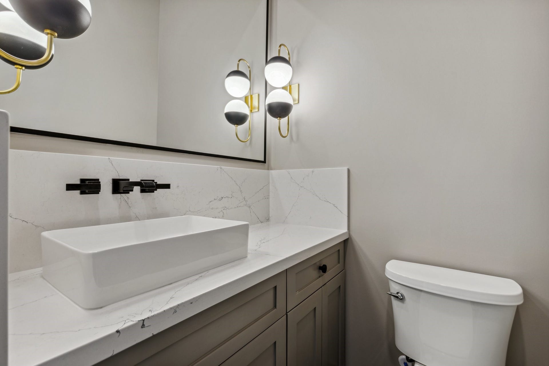 The gorgeous powder room features art deco-inspired tile flooring (not shown), an overmount vessel sink with wall-mounted faucet, and a custom-built vanity. Framed mirror and twin Visual Comfort sconces complete this chic, statement-making space.