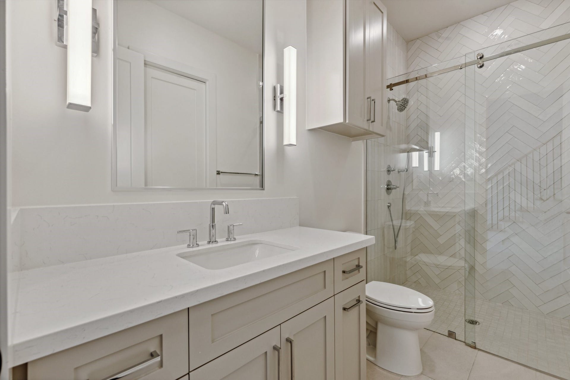 This sleek en-suite bathroom for secondary bedroom #2 features a custom-built vanity with ample storage, Top Knob hardware, and a quartz countertop with an 8-inch backsplash. A framed metal mirror adds a modern touch, while the walk-in shower with herringbone tile, soap niche, and water-repellent sliding glass door elevates the space. Kohler toilet and 24x48 tile flooring complete the clean, contemporary design.