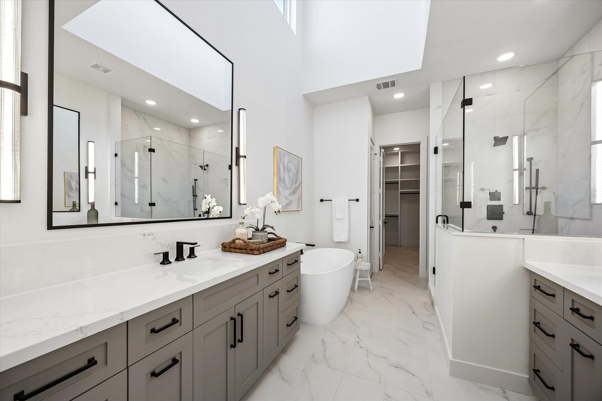 Indulge in a spa-like experience in the primary bath, featuring soaring ceilings, ample natural light from a large window, and double custom-built vanities with Kohler undermount sinks. Visual Comfort sconces frame the mirrors, and sleek porcelain tile floors create a chic, modern look. Straight ahead the door opens to a huge walk-in closet with his and hers dressers and hidden storage that completes the ultimate retreat.