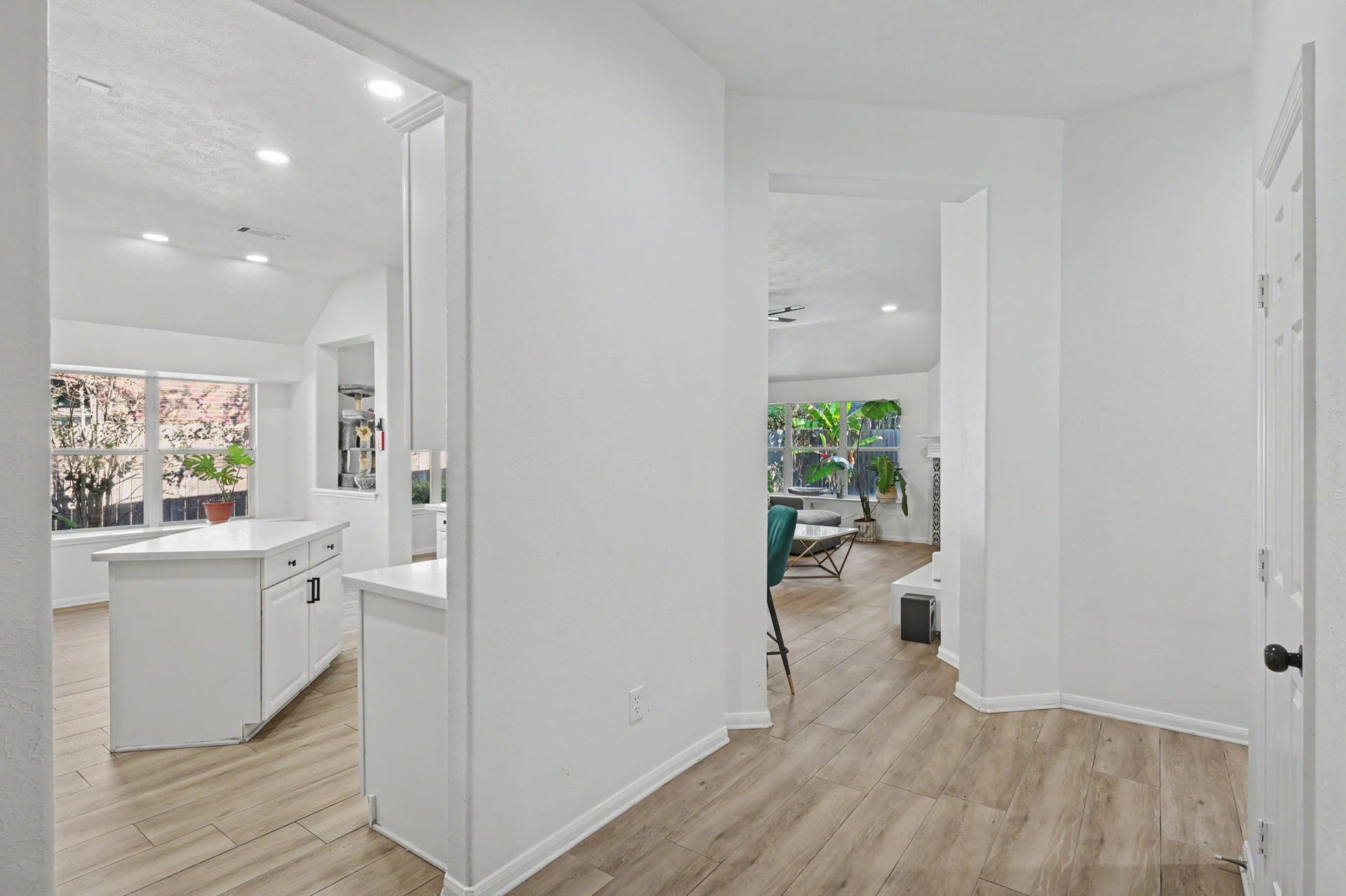 Bright and open hallway leading to a modern kitchen with an island and a living area featuring large windows and wood-style flooring.