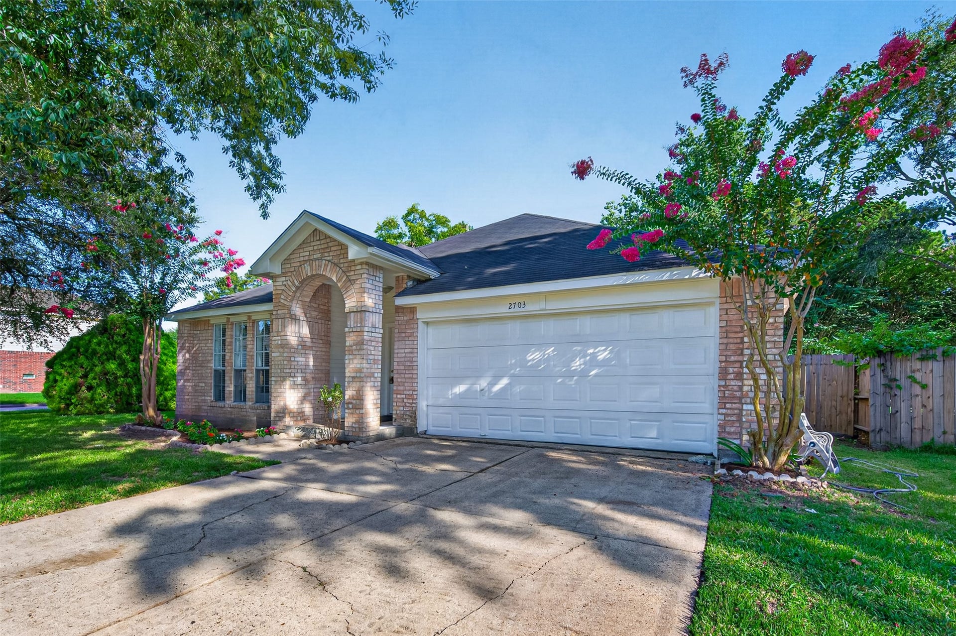 2703 Blue Mist Drive, Sugar Land, TX 77498 Charming single-story brick home with a welcoming front archway, nestled among mature trees and vibrant landscaping, featuring a spacious driveway and a two-car garage.