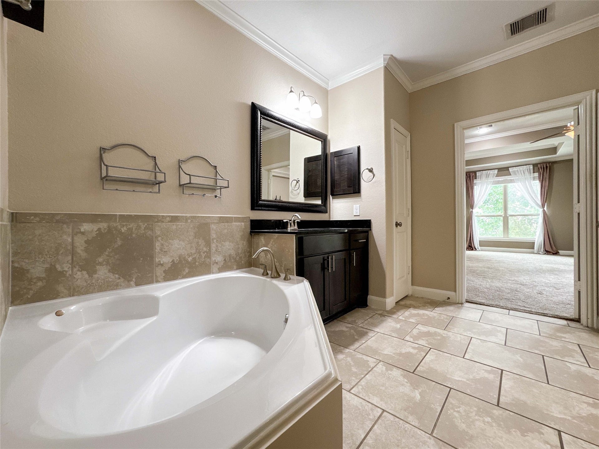 This is a spacious bathroom featuring a large soaking tub, a sleek vanity with a framed mirror, and neutral tile flooring. It opens to a bright bedroom with carpet and large windows, creating a seamless flow between the spaces.