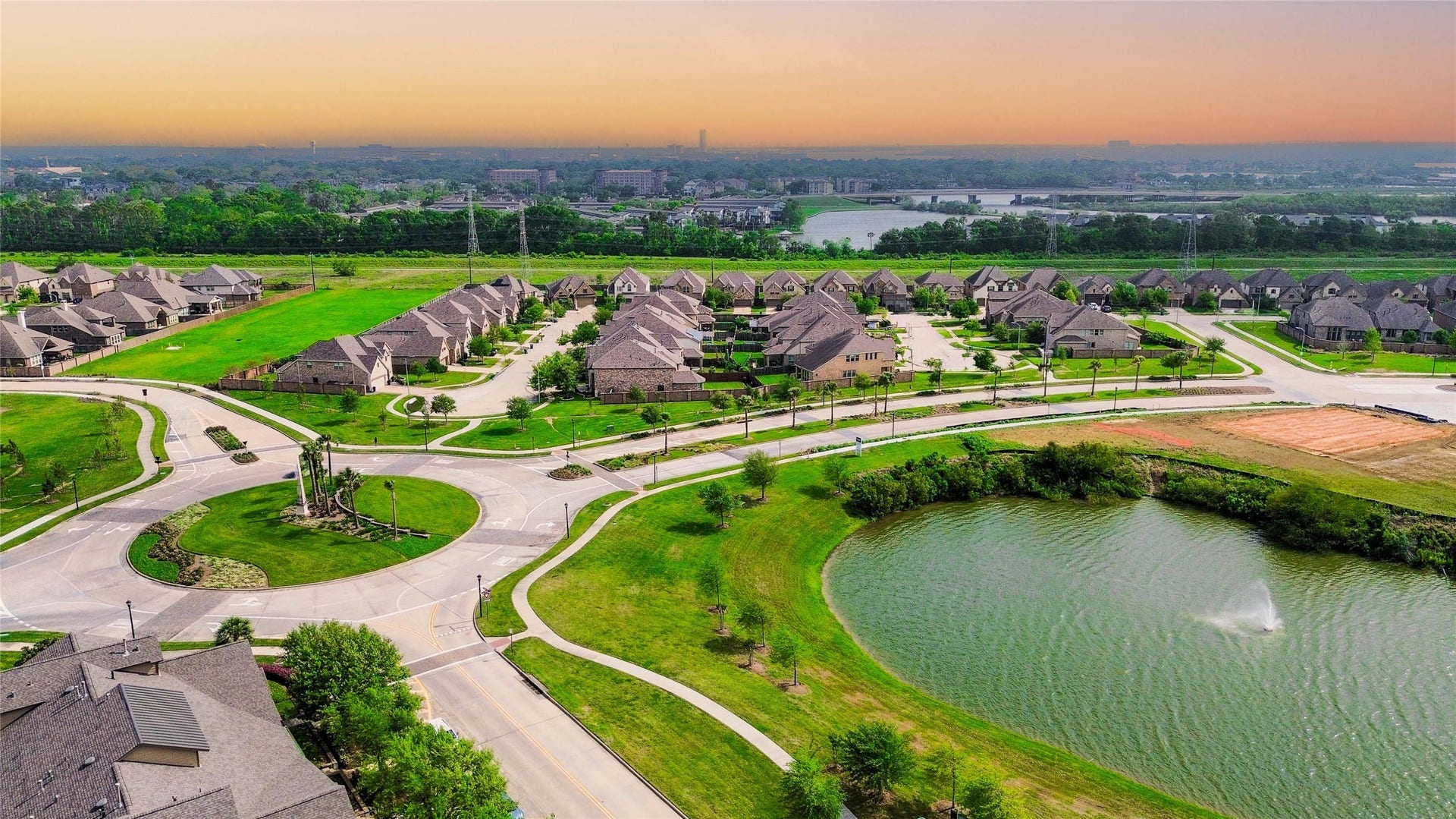 This aerial photo showcases a suburban neighborhood with modern homes, winding roads, and ample green spaces. A scenic pond with a fountain adds a serene touch, while the expansive view highlights the community's organized layout and proximity to nature.