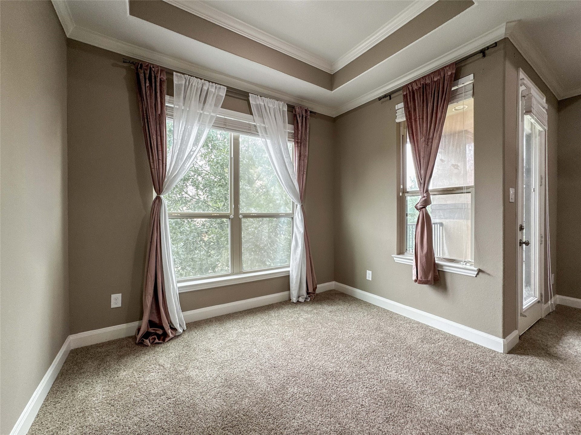 Bright room with large windows, neutral walls, and carpeted floors. Features a tray ceiling and elegant drapery, creating a cozy atmosphere.
