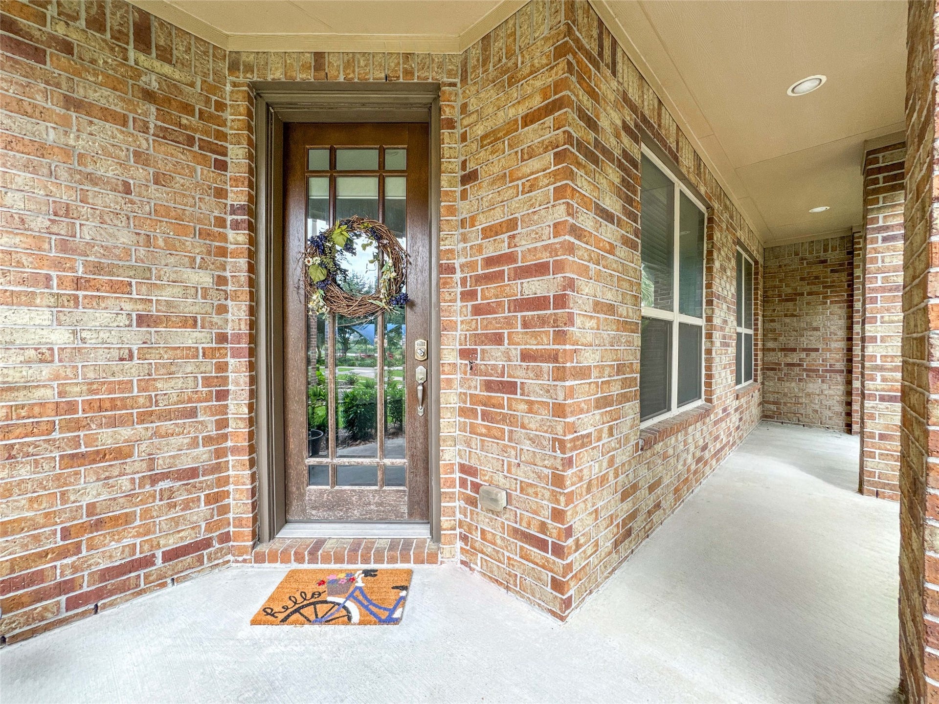 This photo showcases a welcoming brick entryway with a charming wooden front door, adorned with a decorative wreath and a colorful welcome mat. The area is well-lit and features a covered porch, offering a cozy and inviting entrance to the home.
