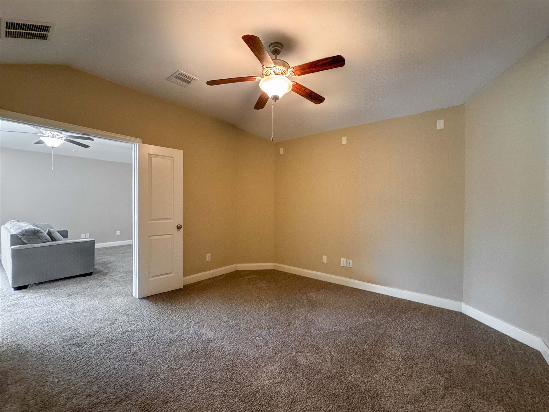 This room features a neutral color palette with carpeted flooring, a ceiling fan, and ample natural light. It connects to another room, offering flexibility for use as a bedroom or office space.
