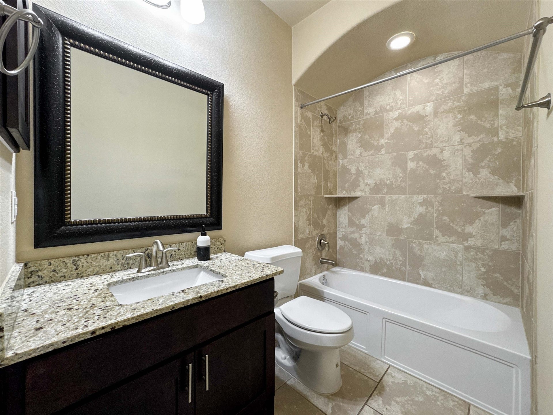 This bathroom features a modern design with a granite countertop, dark wood vanity, and a large framed mirror. It includes a bathtub with a tiled shower area and recessed lighting for a clean, elegant look.