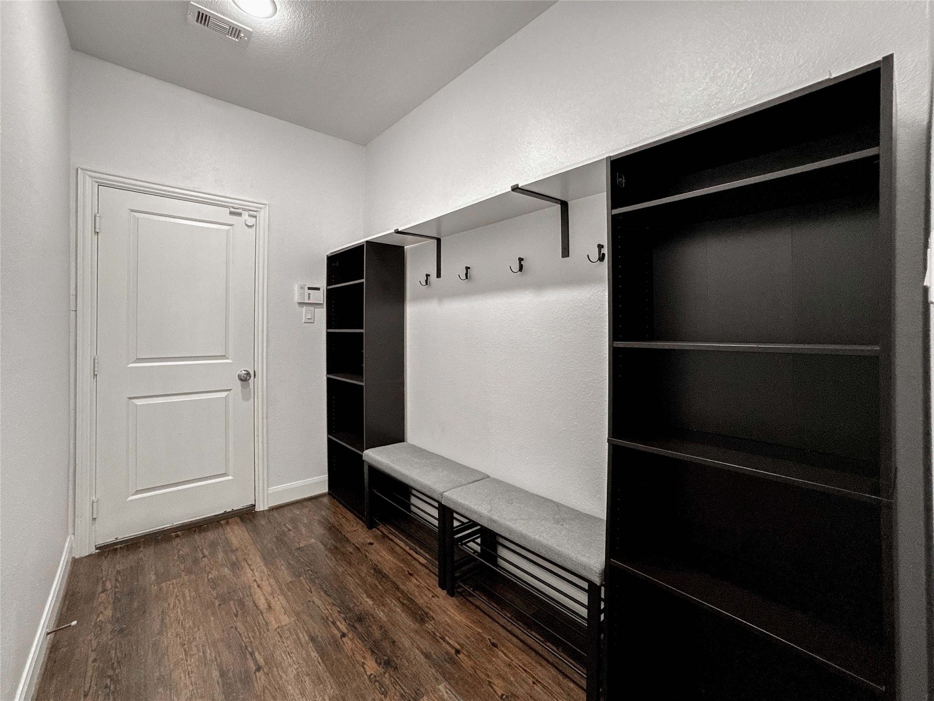 Stay organized with this mudroom with luxury vinyl flooring, white walls, and a door leading to the garage. It features black shelving units, a bench with hooks above for hanging, and overhead lighting.