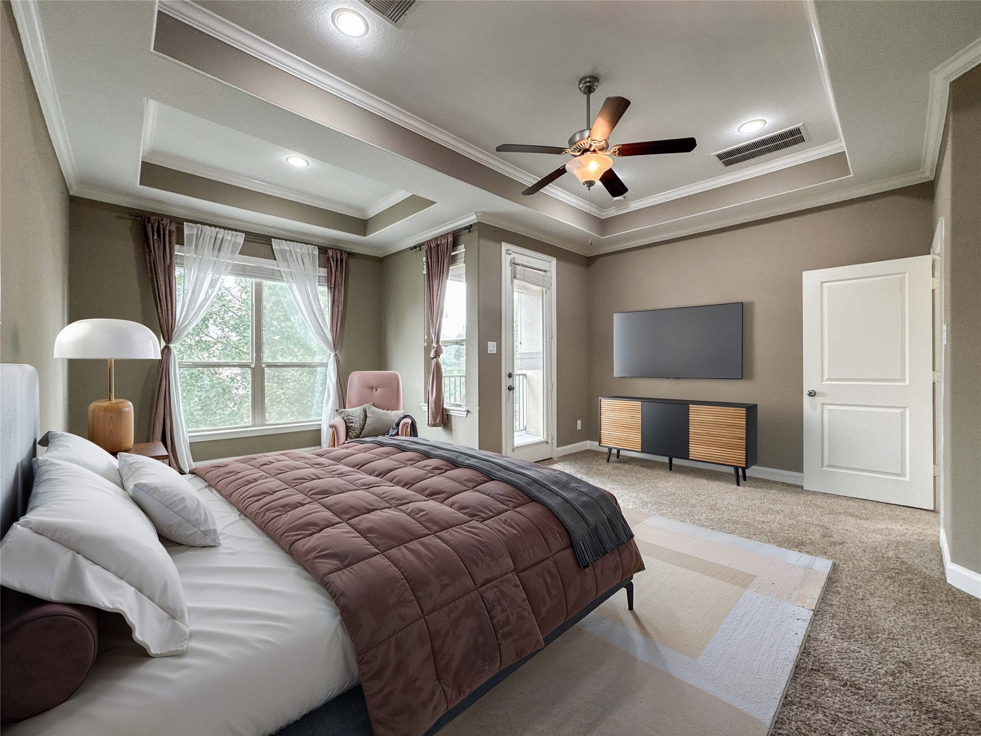The primary bedroom features a tray ceiling with recessed lighting and a ceiling fan. It has large windows with elegant curtains, a cozy carpet, and ample space for a TV setup. The room is inviting, well-lit, and perfect for relaxation.